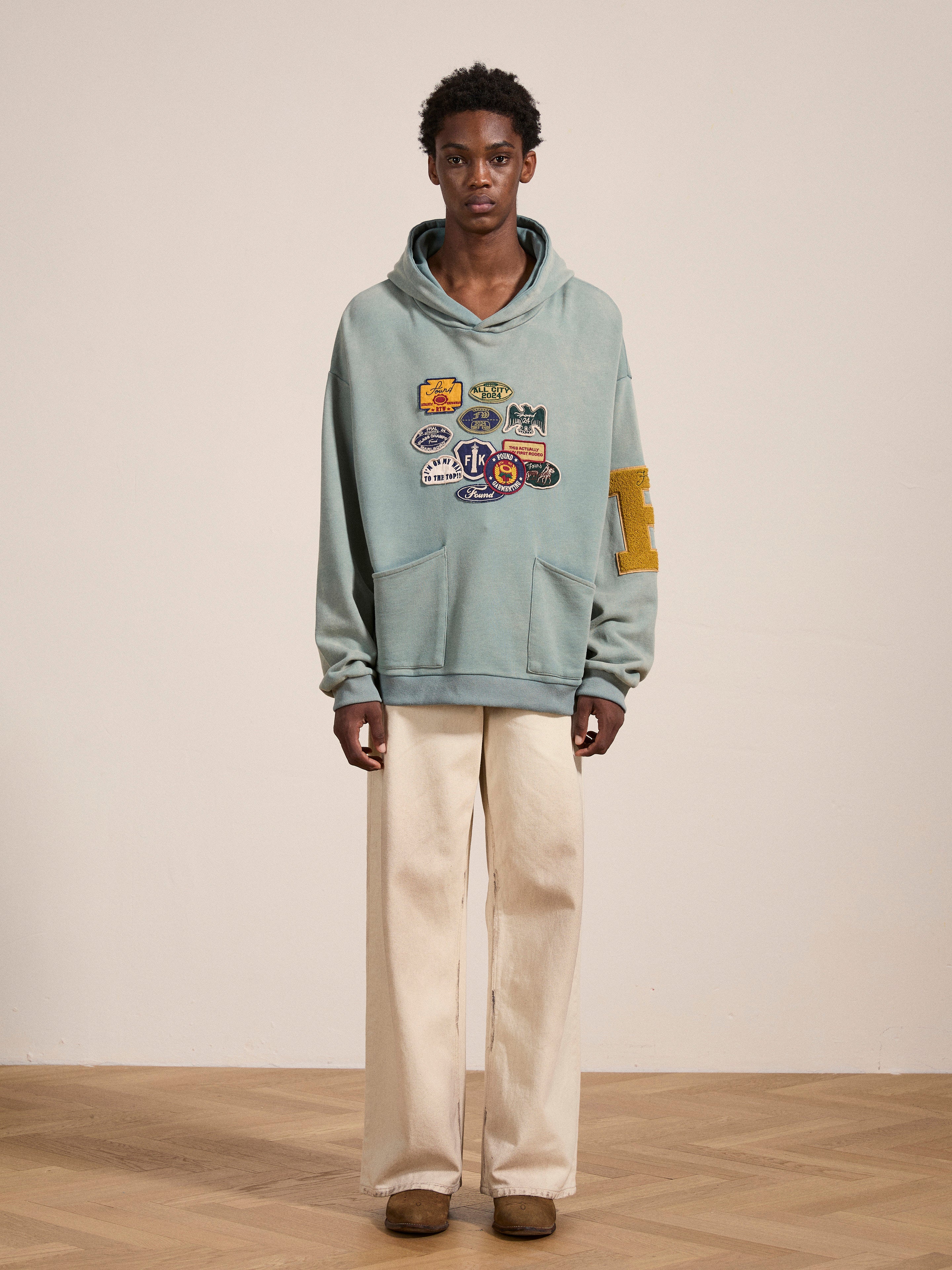 A person standing indoors, wearing FOUND's Multi Patch Hoodie crafted from vintage-inspired French Terry cotton in sage green, adorned with multiple patches on the front and a large letter "E" on the sleeve, paired with off-white pants and brown shoes.