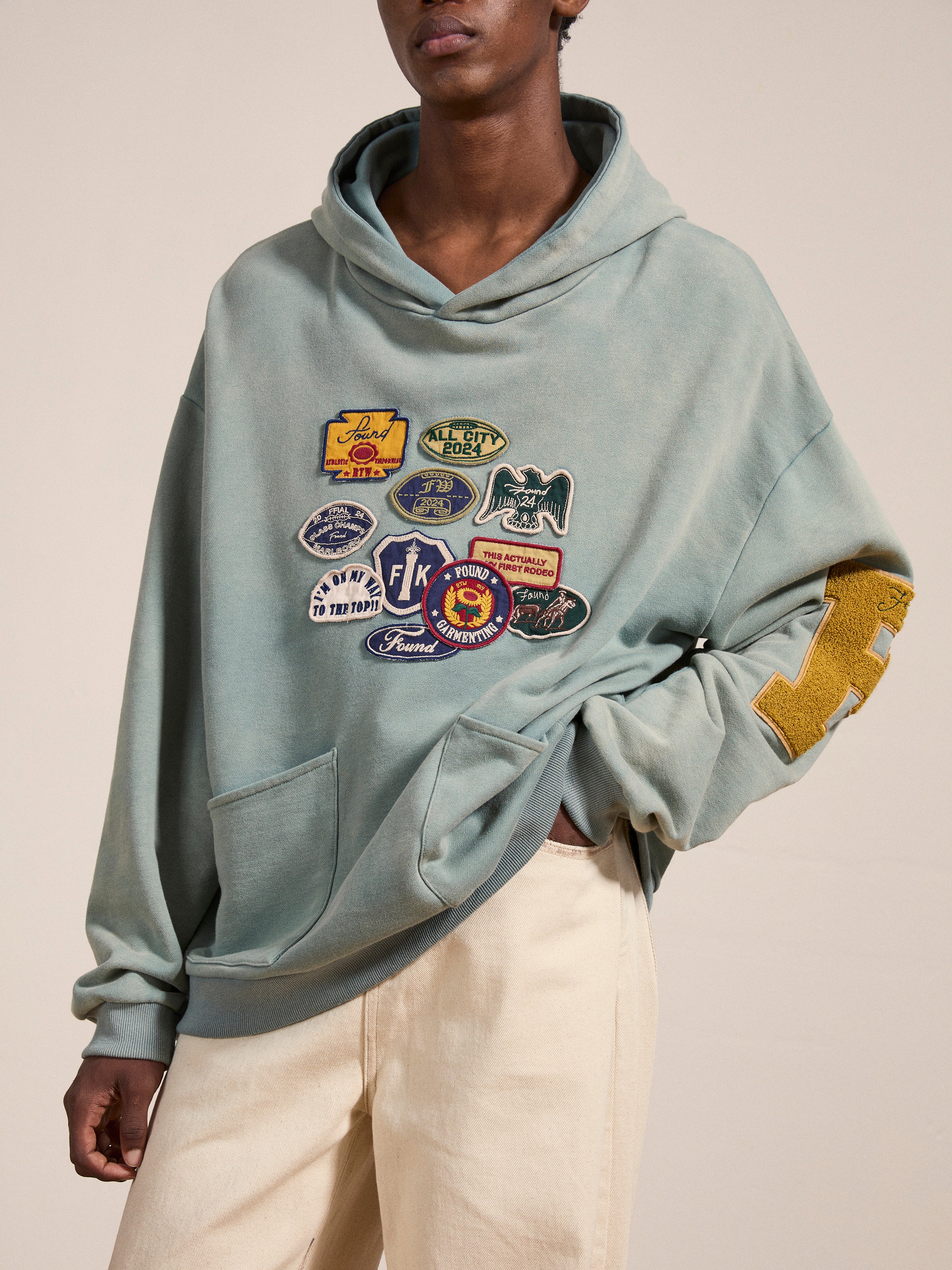 Person wearing a vintage-inspired Multi Patch Hoodie from FOUND, made from French Terry cotton, featuring various patches on the front and a yellow letter patch on the sleeve. They are also wearing beige pants.