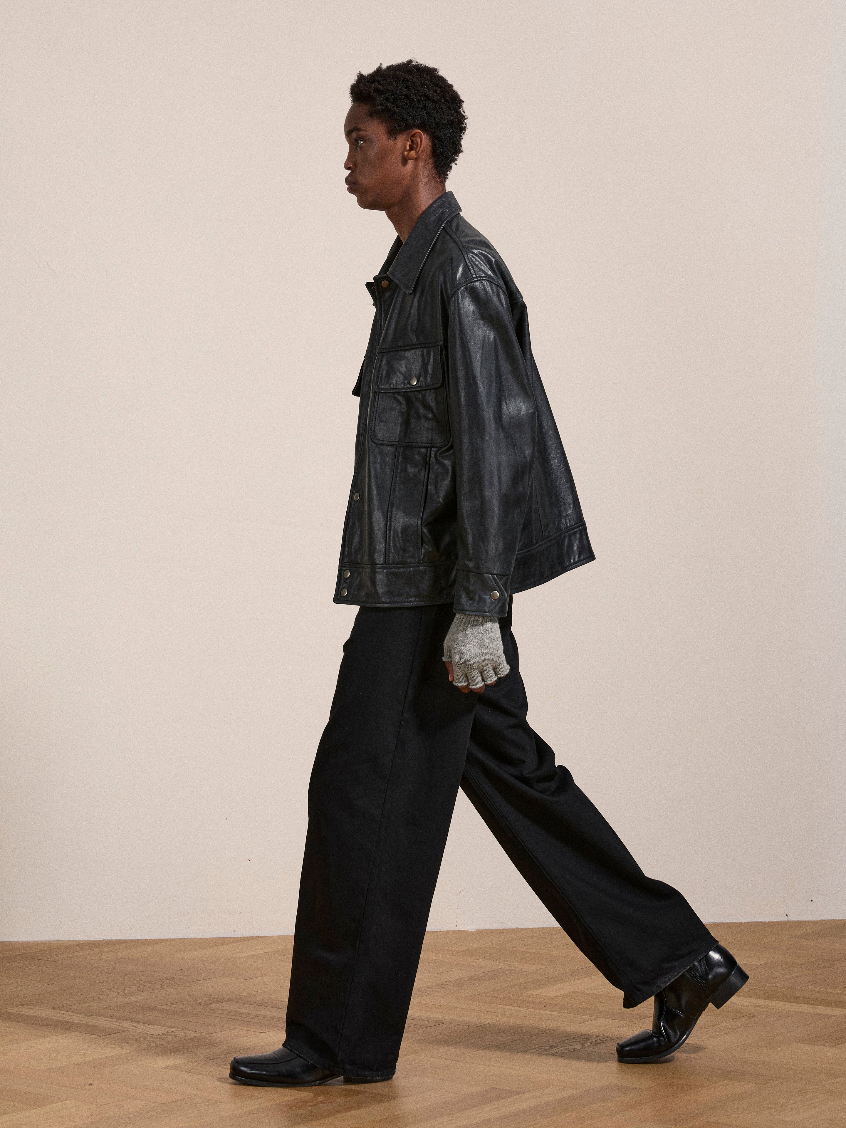 A person is walking indoors, wearing a black FOUND Leather Trucker Jacket, black wide-leg pants, black shoes, and gray gloves. The background features a plain light-colored wall and wooden floor.