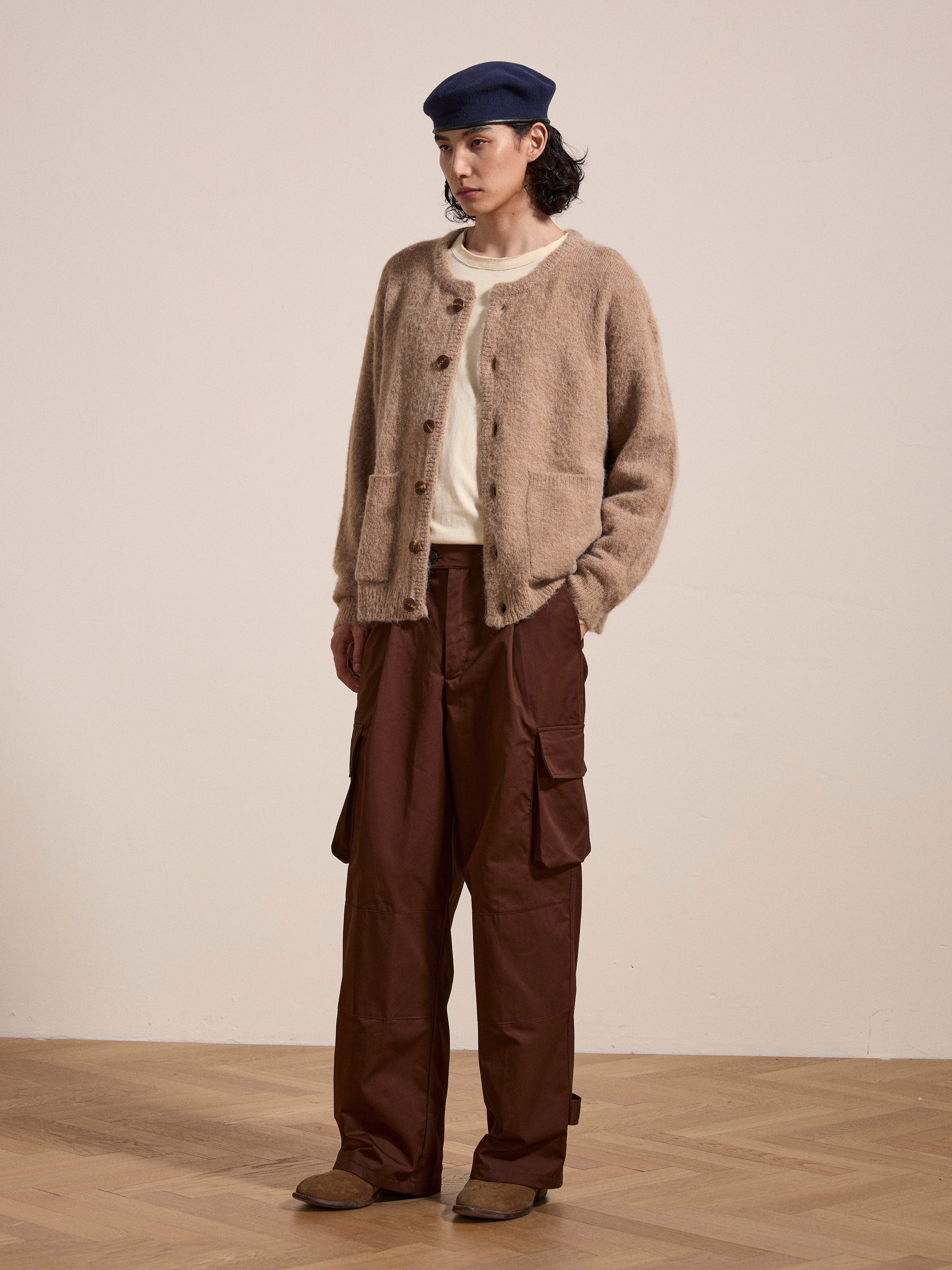 A person stands on a wooden floor wearing an oversized fit Mohair Cardigan in beige with tortoise buttons from FOUND, a white shirt, brown cargo pants, a navy beret, and brown shoes. The background is plain white.