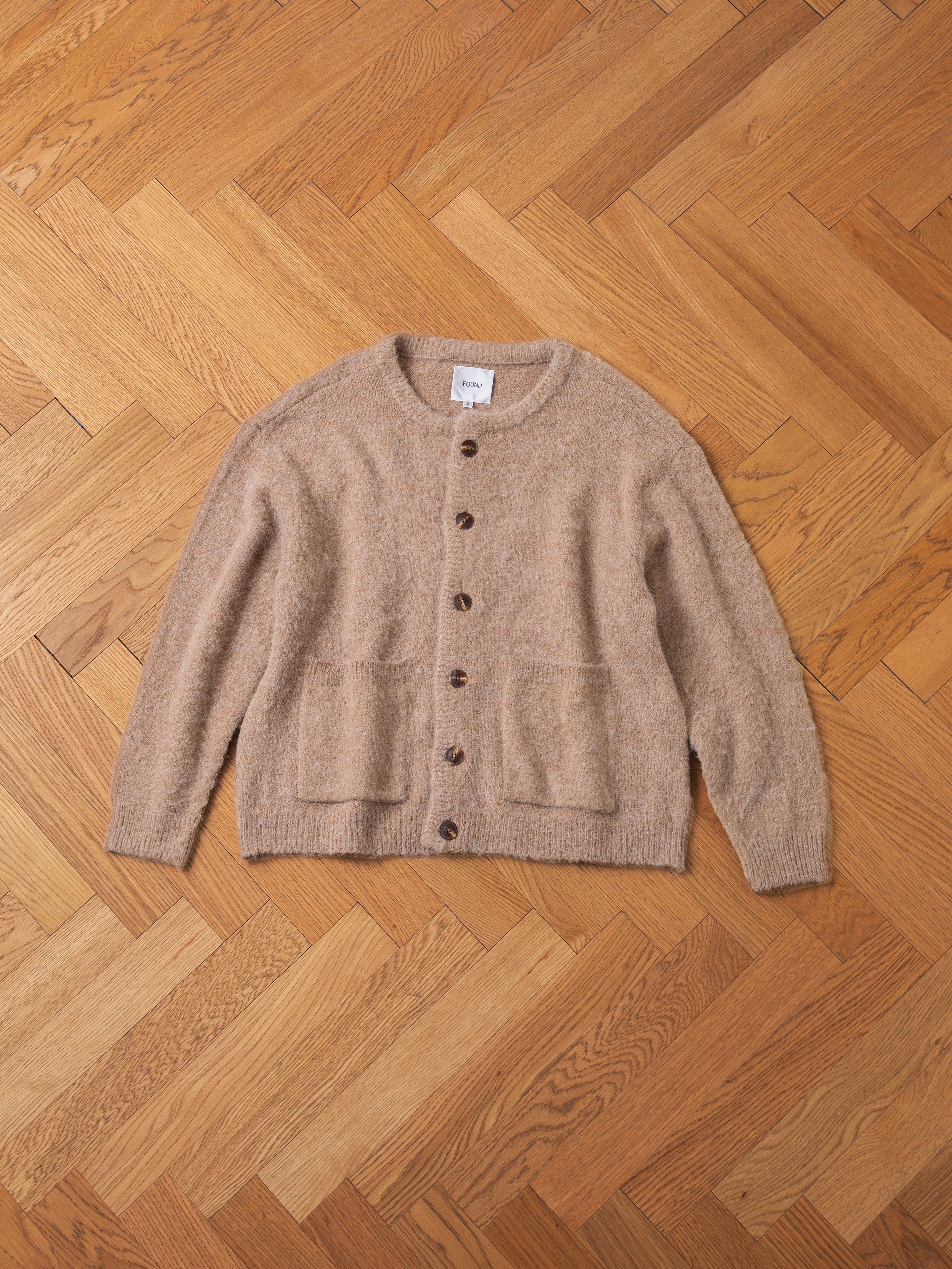 The FOUND Mohair Cardigan, featuring an oversized fit in beige with tortoise buttons and two front pockets, is laid flat on a wooden parquet floor.