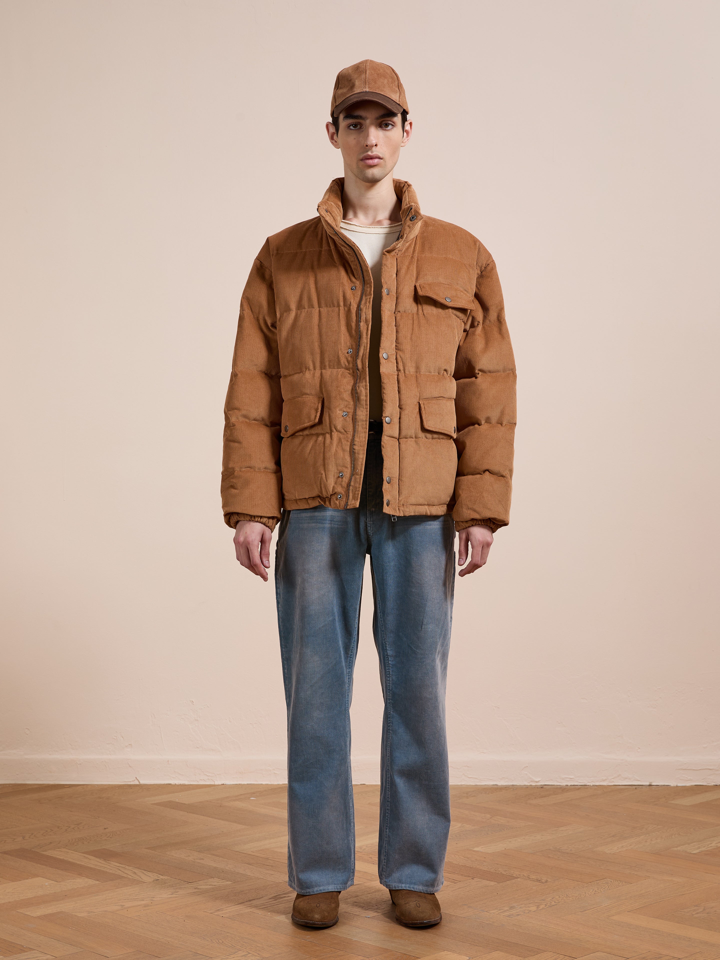 A person stands indoors against a plain background, wearing a relaxed fit FOUND Corduroy Puffer Jacket in brown, styled with a brown cap, white shirt, loose blue jeans, and brown shoes.