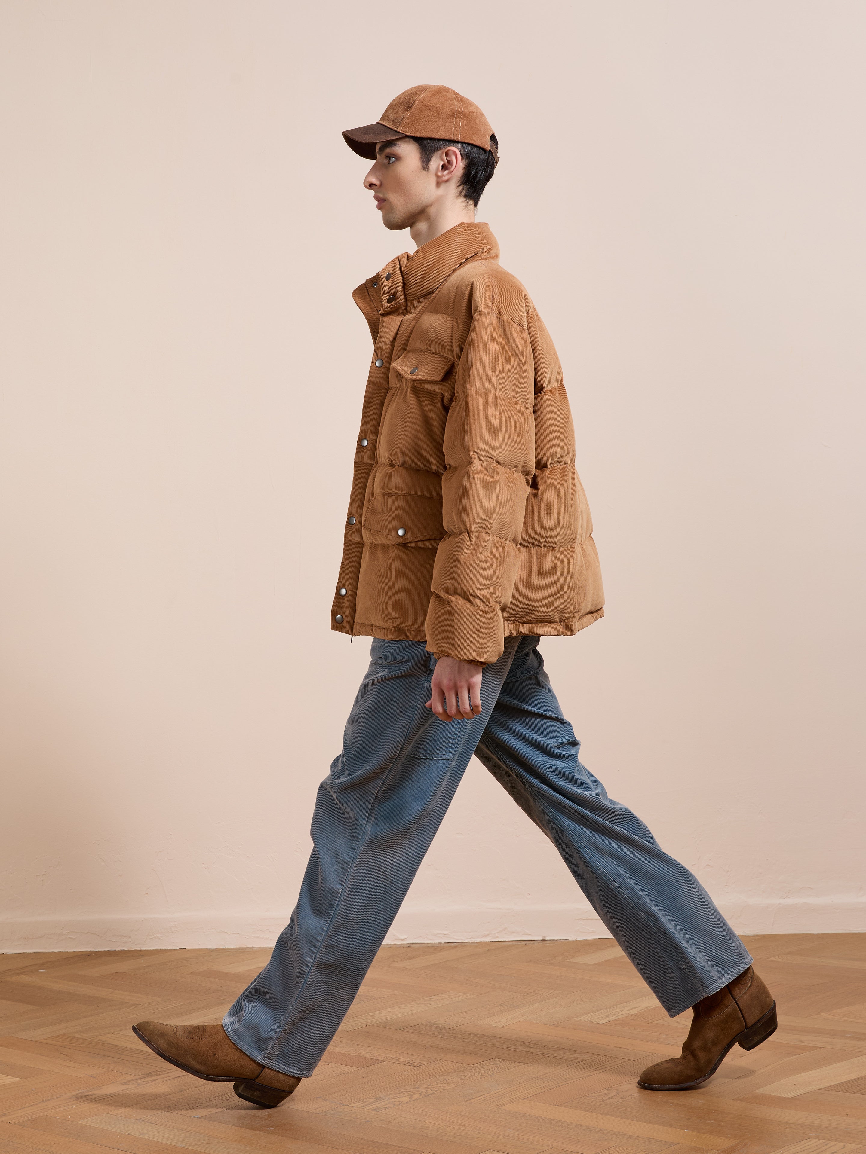 A person wearing FOUND’s Corduroy Puffer Jacket in brown, with a matching brown cap, blue jeans, and brown boots walks indoors on a wooden floor against a plain light-colored wall.