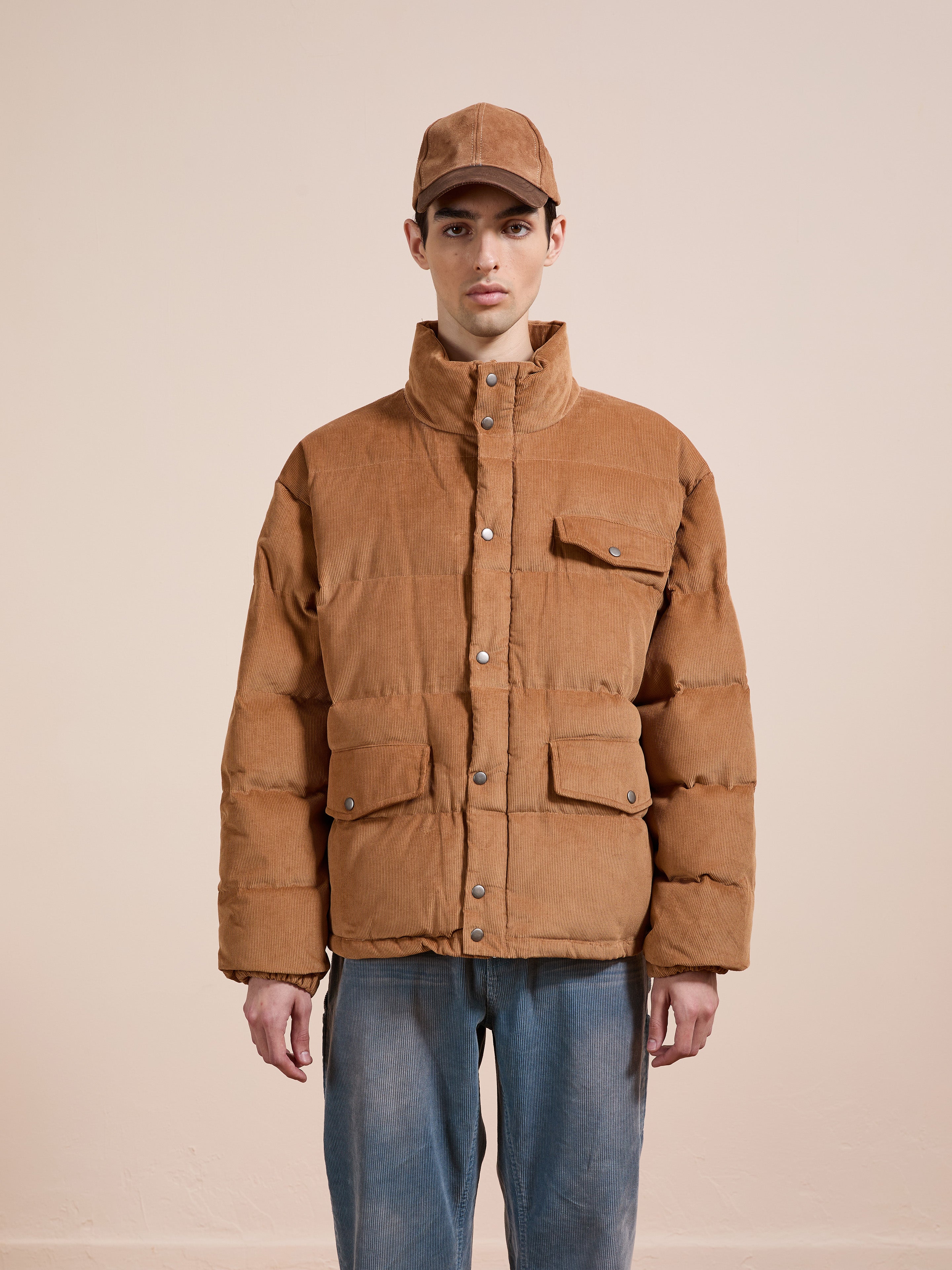 A person wears the FOUND Corduroy Puffer Jacket and a brown cap with blue jeans, standing facing forward against a plain beige background.