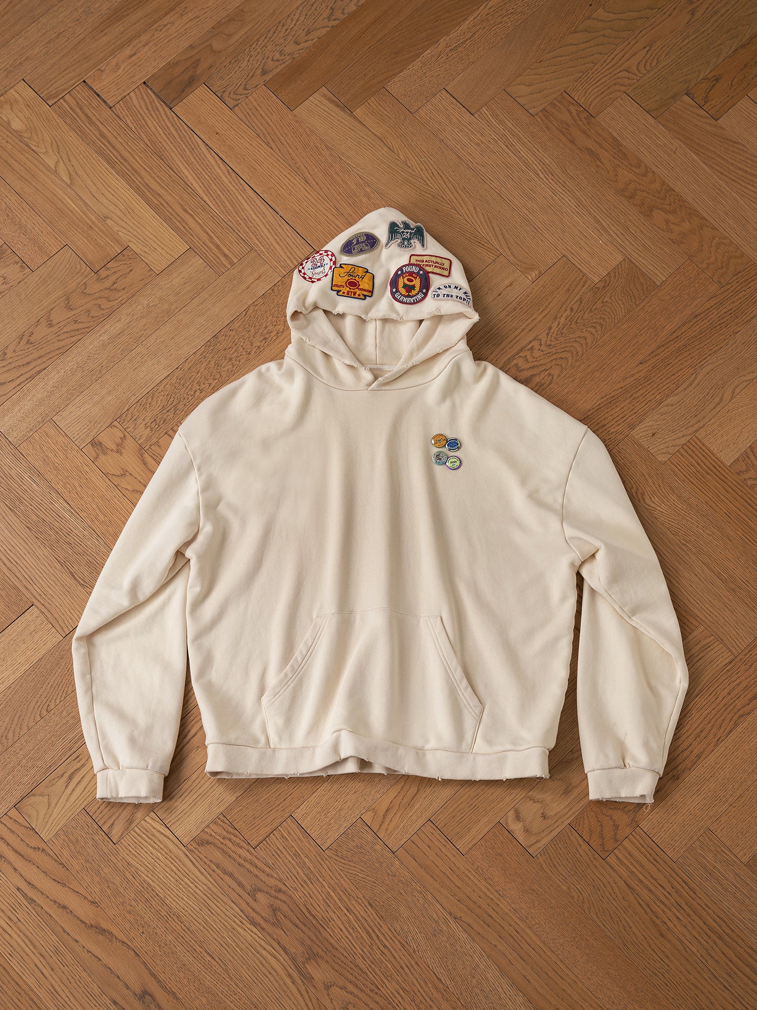 FOUND's Faded Patch Hoodie showcases a vintage flair with its oversized fit, embellished with an array of patches on the hood and chest, elegantly displayed flat on a wooden floor.
