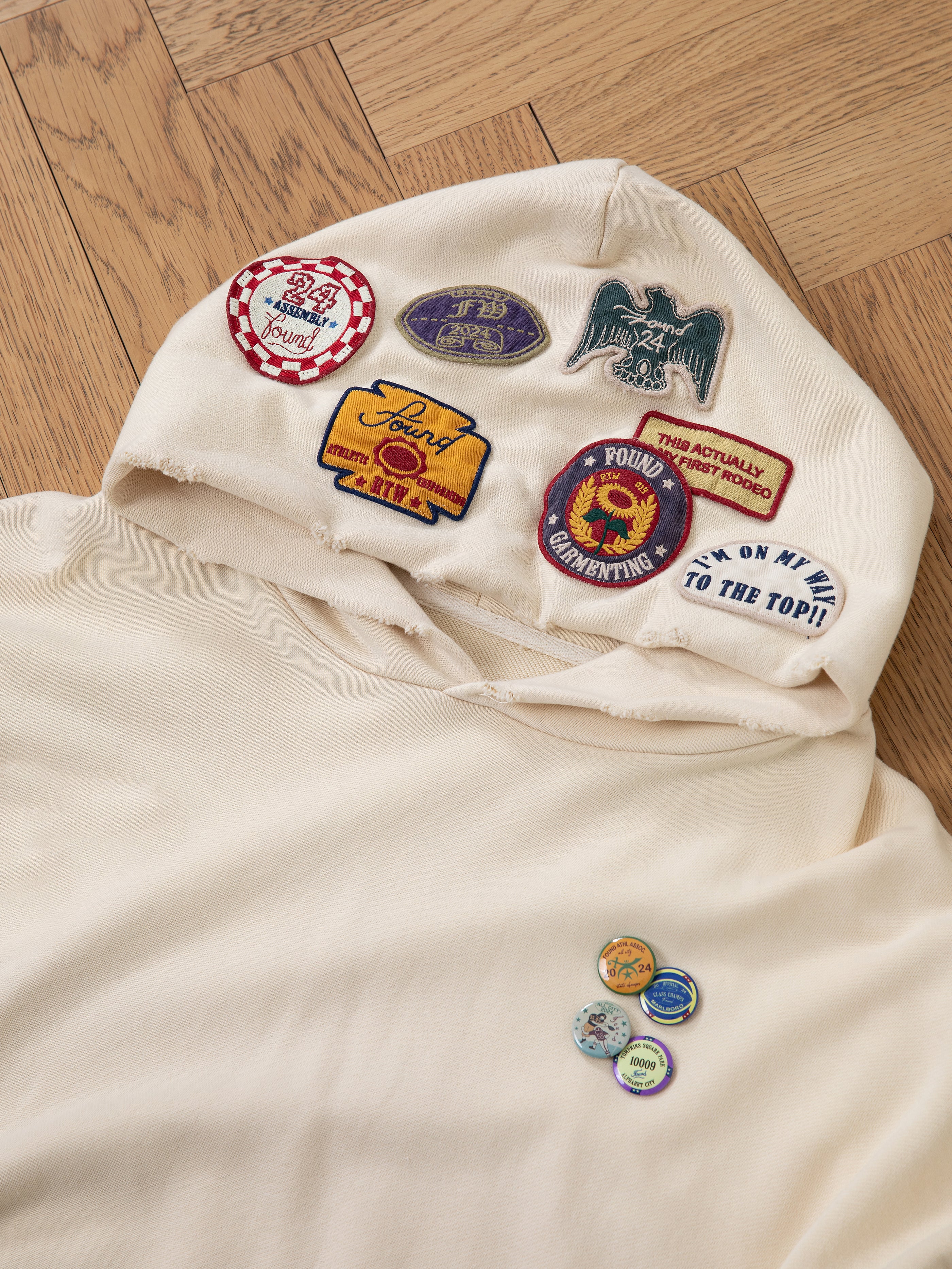 The Faded Patch Hoodie by FOUND, in a cream color with an oversized fit, showcases a variety of colorful embroidered patches on the hood and near the pocket. Placed on a wooden floor, its vintage style brings a touch of timeless charm.