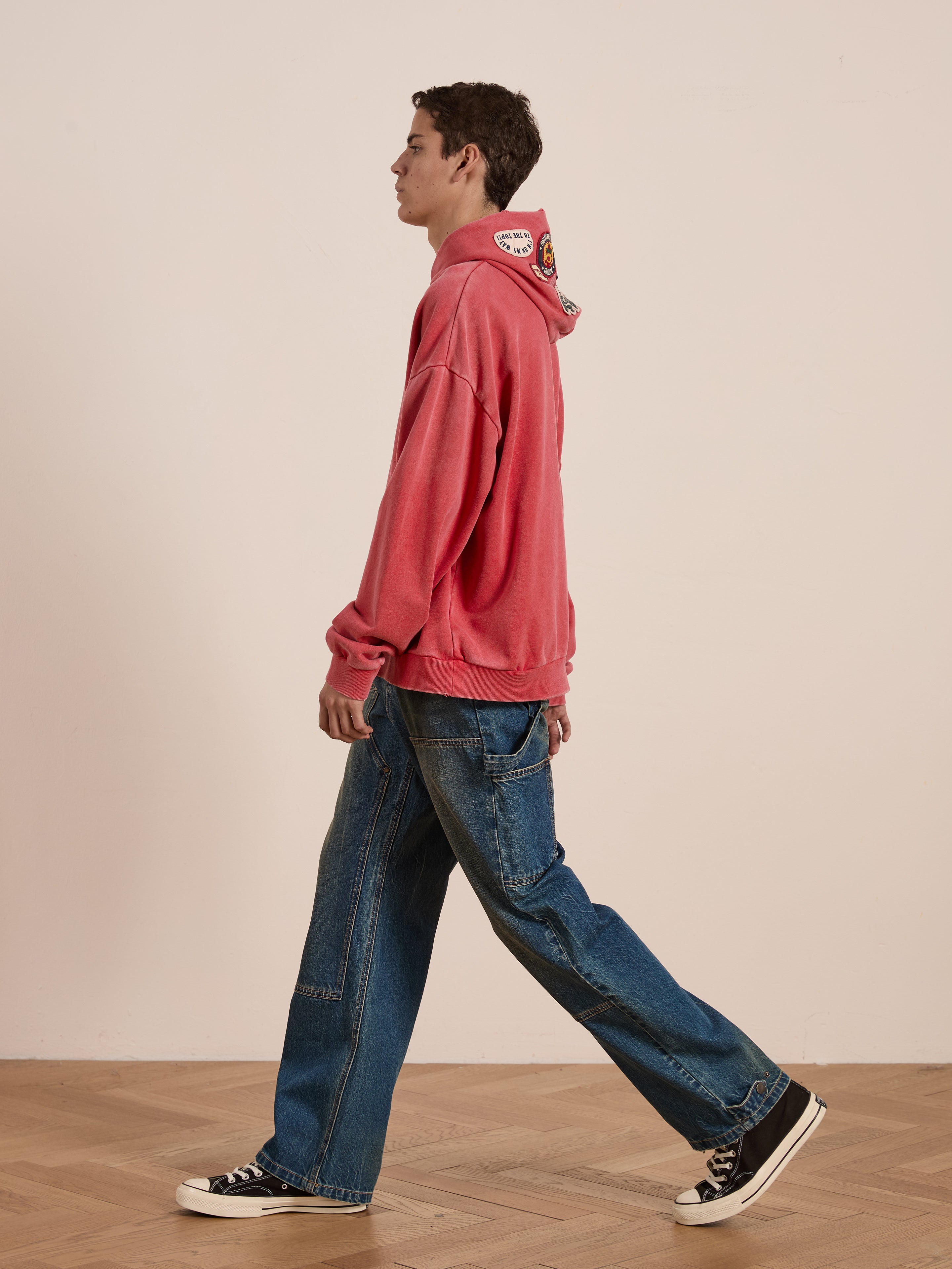A person wearing a red Multi Patch Hoodie by FOUND with blue distressed jeans walks indoors on a vintage-inspired wooden floor, facing sideways.