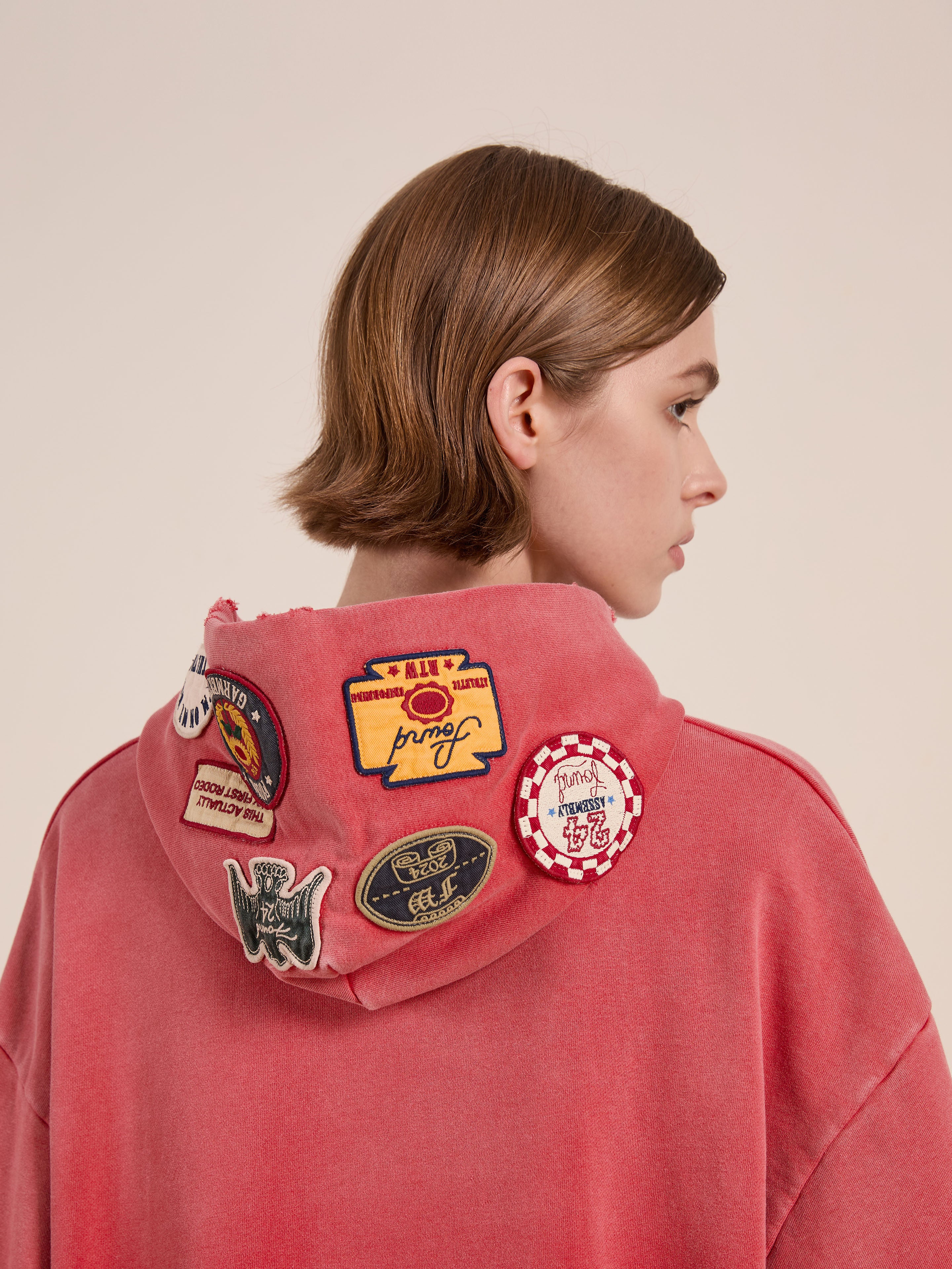 A person in a FOUND Multi Patch Hoodie, featuring colorful patches on the hood's back and vintage-inspired design, stands sideways against a plain backdrop. Its unisex charm offers an effortlessly timeless appeal.