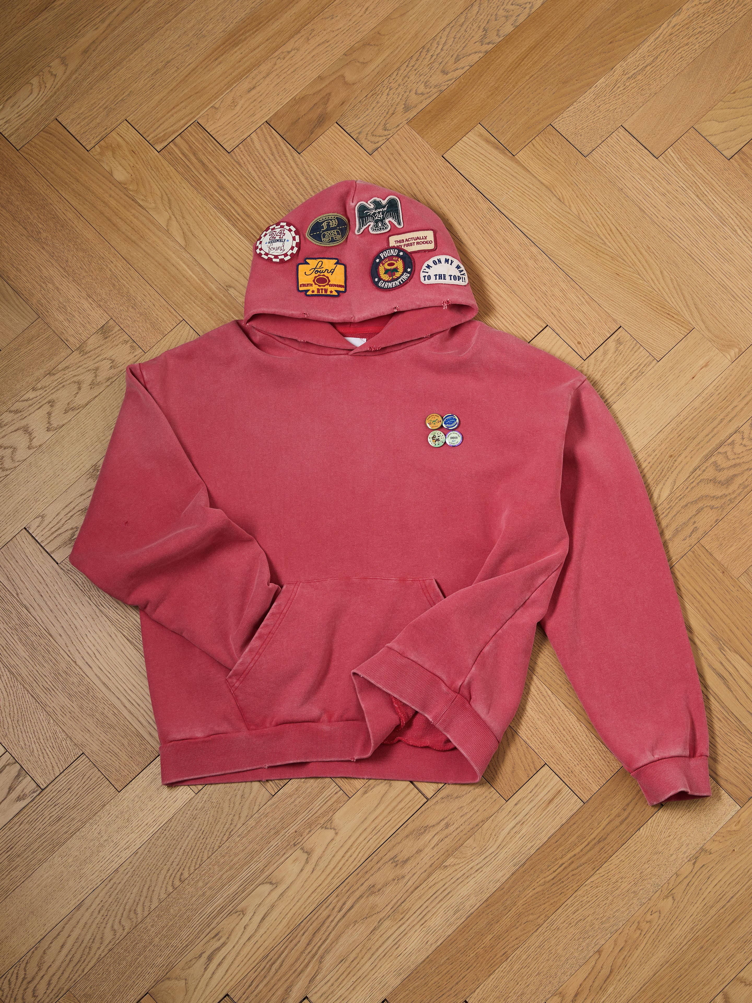 The FOUND Multi Patch Hoodie, featuring a sun-faded red wash with colorful patches and badges, is displayed on a wooden herringbone floor.