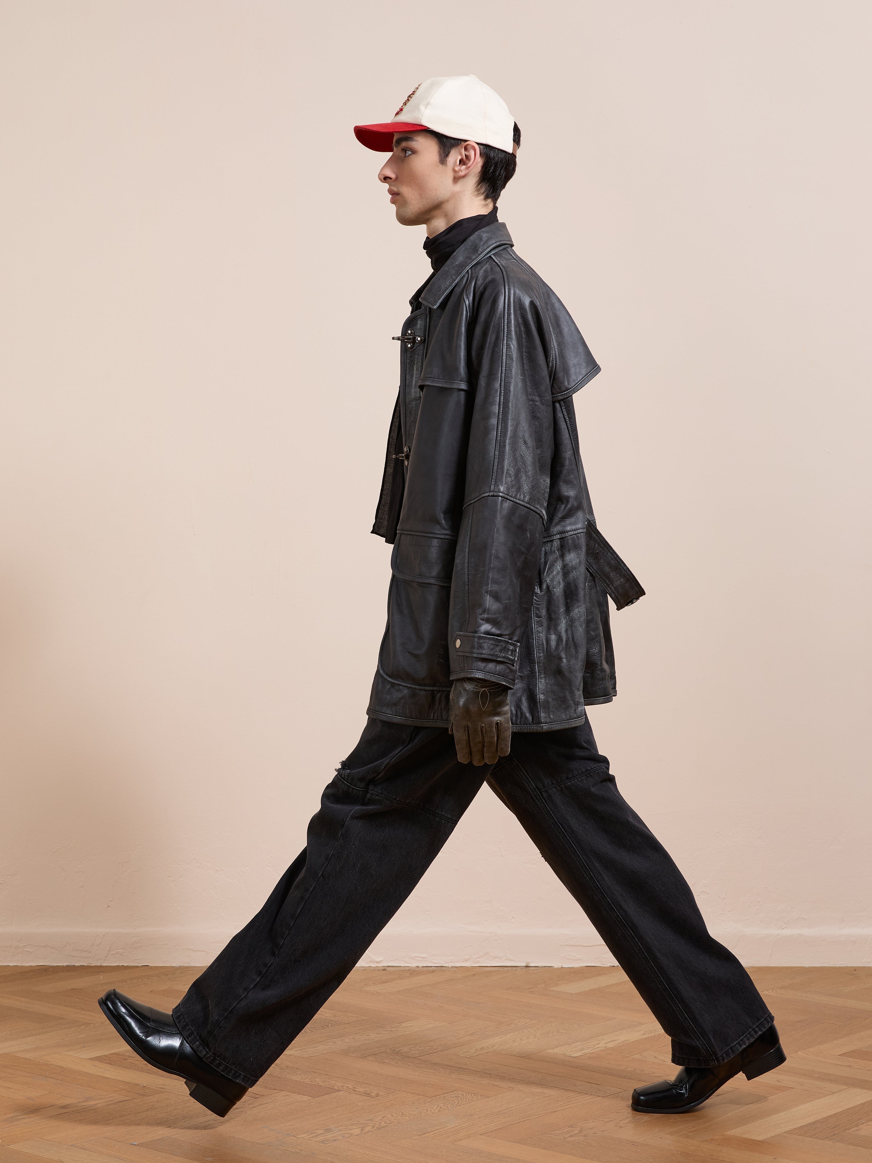 A person wearing the FOUND Distressed Leather Fireman Clasp Jacket, a white and red cap, dark pants, and black heeled shoes walks briskly indoors against a plain beige wall.