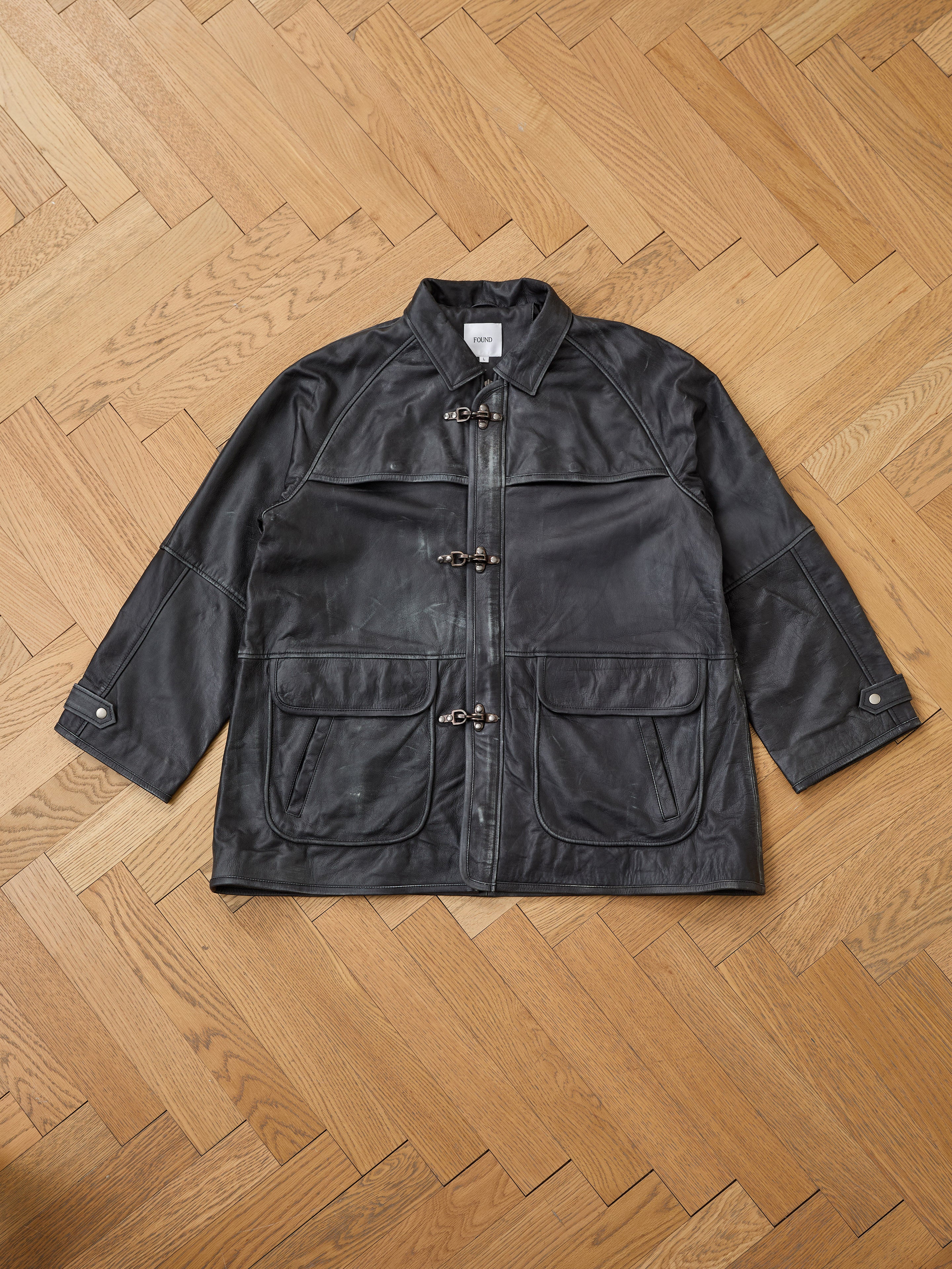 The FOUND Distressed Leather Fireman Clasp Jacket, a unisex genuine leather piece with front zipper, toggle closures, two large pockets, and buttoned cuffs, is laid flat on a parquet floor.