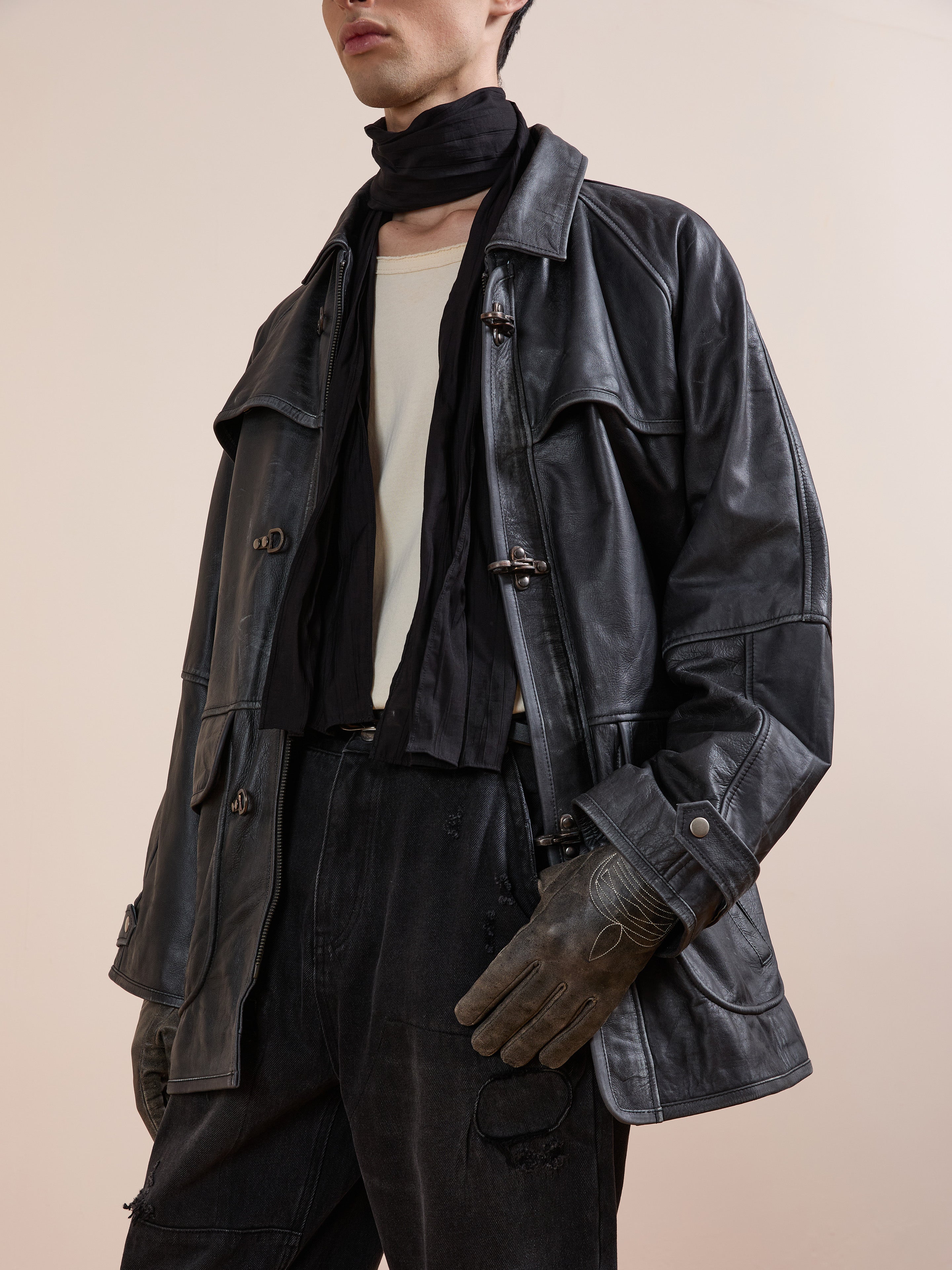 A person wearing the FOUND Distressed Leather Fireman Clasp Jacket, black scarf, distressed black jeans, a white shirt, and brown gloves, posing against a plain beige background.
