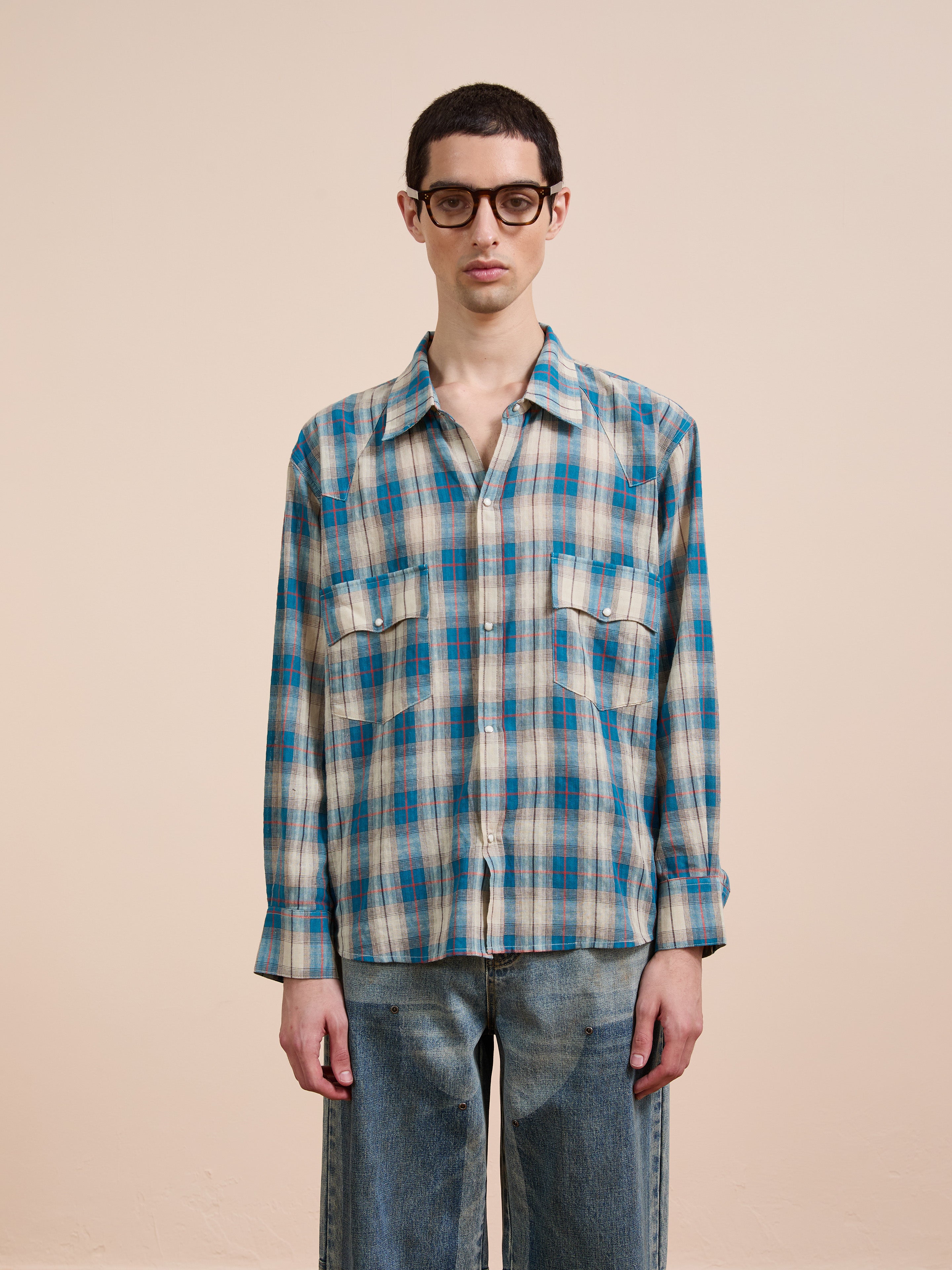 A person wearing glasses, a blue and beige FOUND Flannel Shirt with a relaxed unisex fit, and blue jeans stands against a plain beige background.