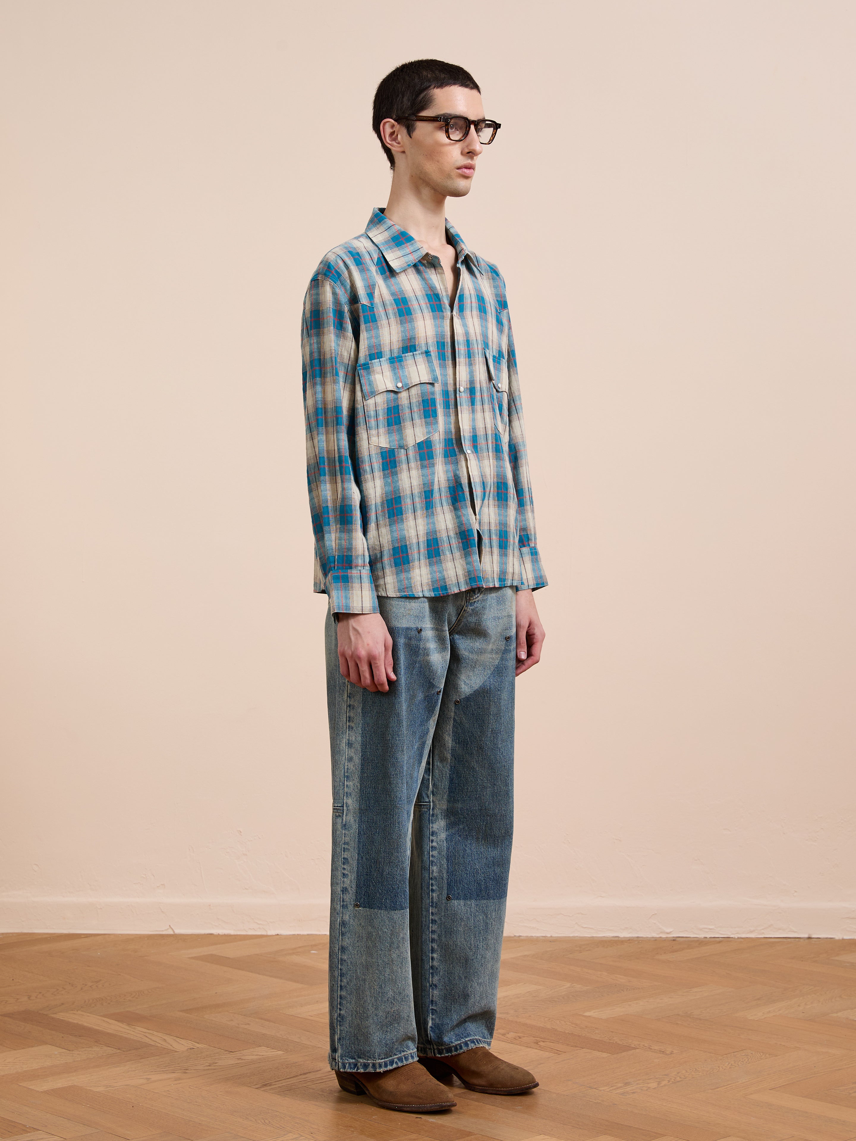 A person stands indoors on a wooden floor, wearing glasses, patchwork jeans, brown shoes, and a FOUND Flannel Shirt in a relaxed unisex fit with a Western plaid pattern, facing sideways against a plain light wall.