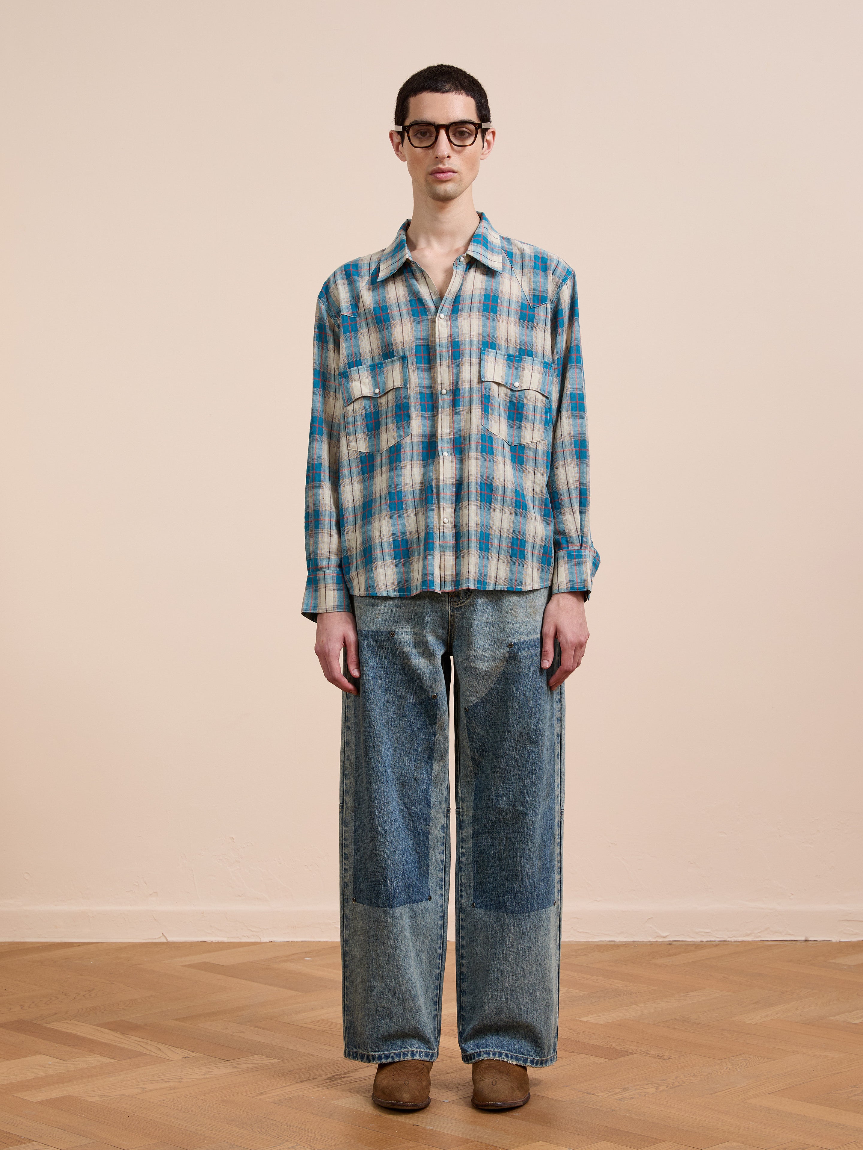 A person wears the FOUND Flannel Shirt with a unisex relaxed fit, wide-leg patched jeans, black glasses, and brown shoes, standing on a wooden floor against a plain beige wall.
