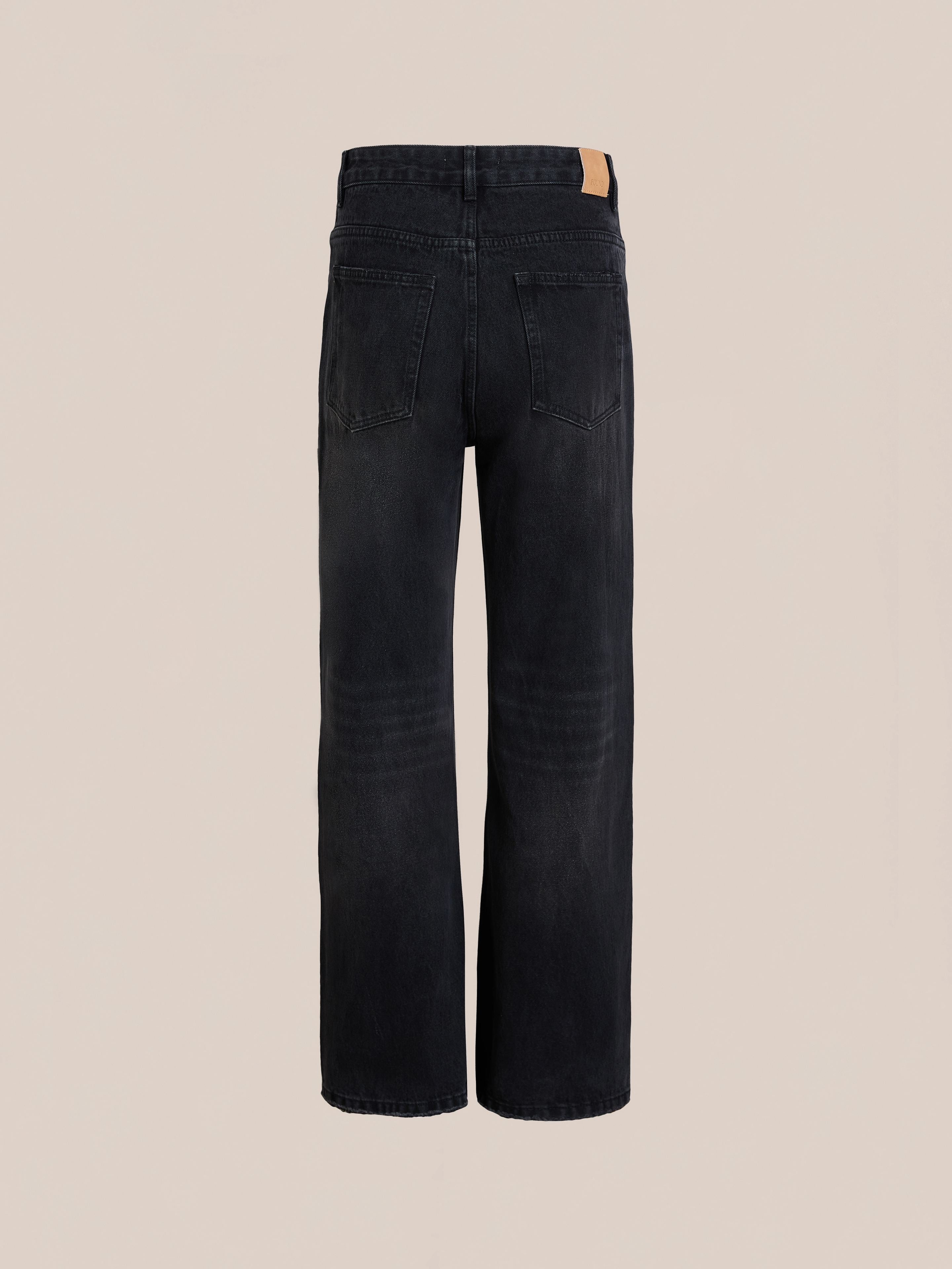 Back view of Found Flare Jeans in vintage black wash featuring two rear pockets, a tan leather patch on the waistband, and subtly distressed hems, displayed against a plain beige background.