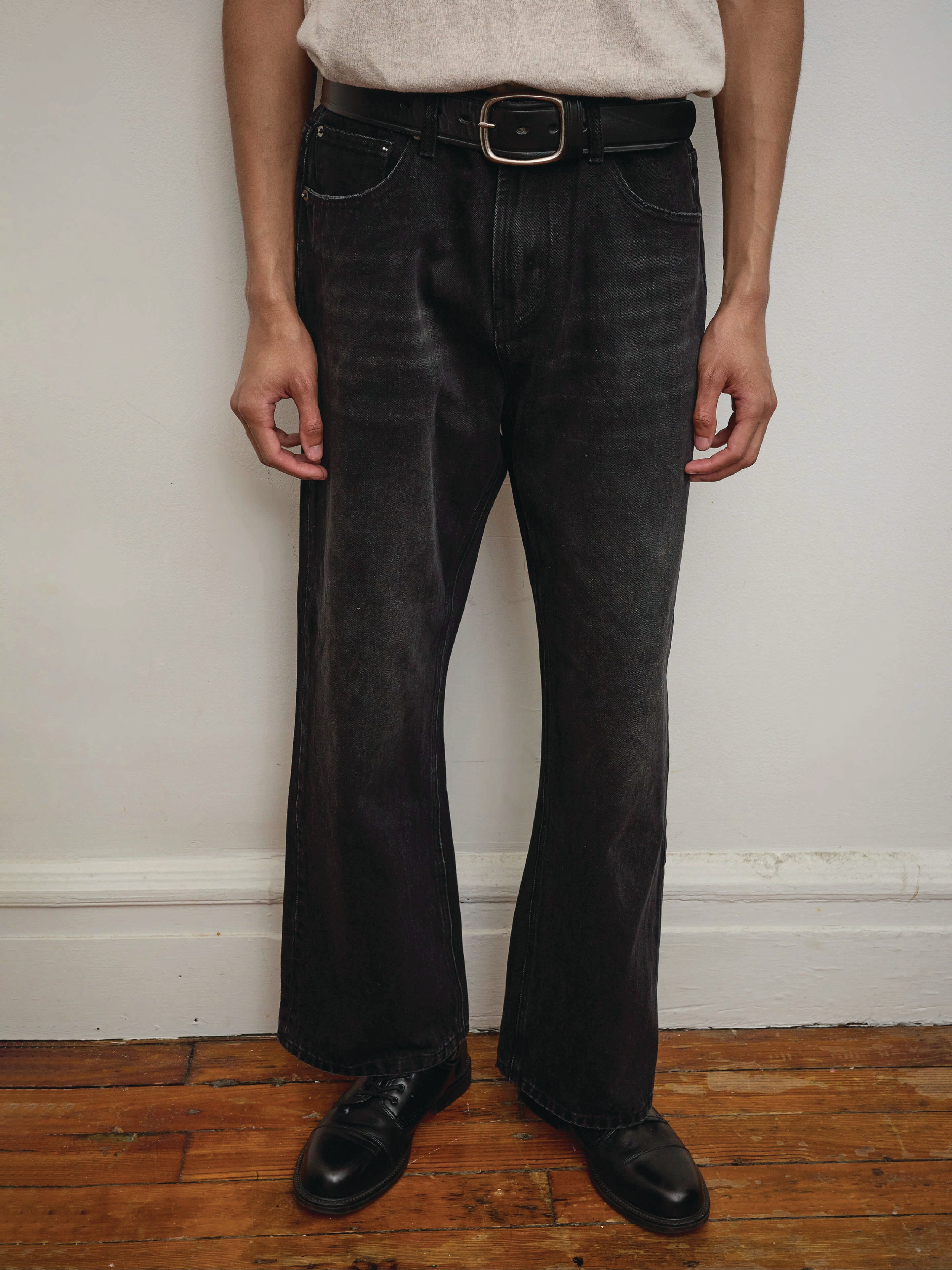 A person wearing Found Flare Jeans, a black belt, black shoes, and a beige shirt stands against a plain wall on a wooden floor.