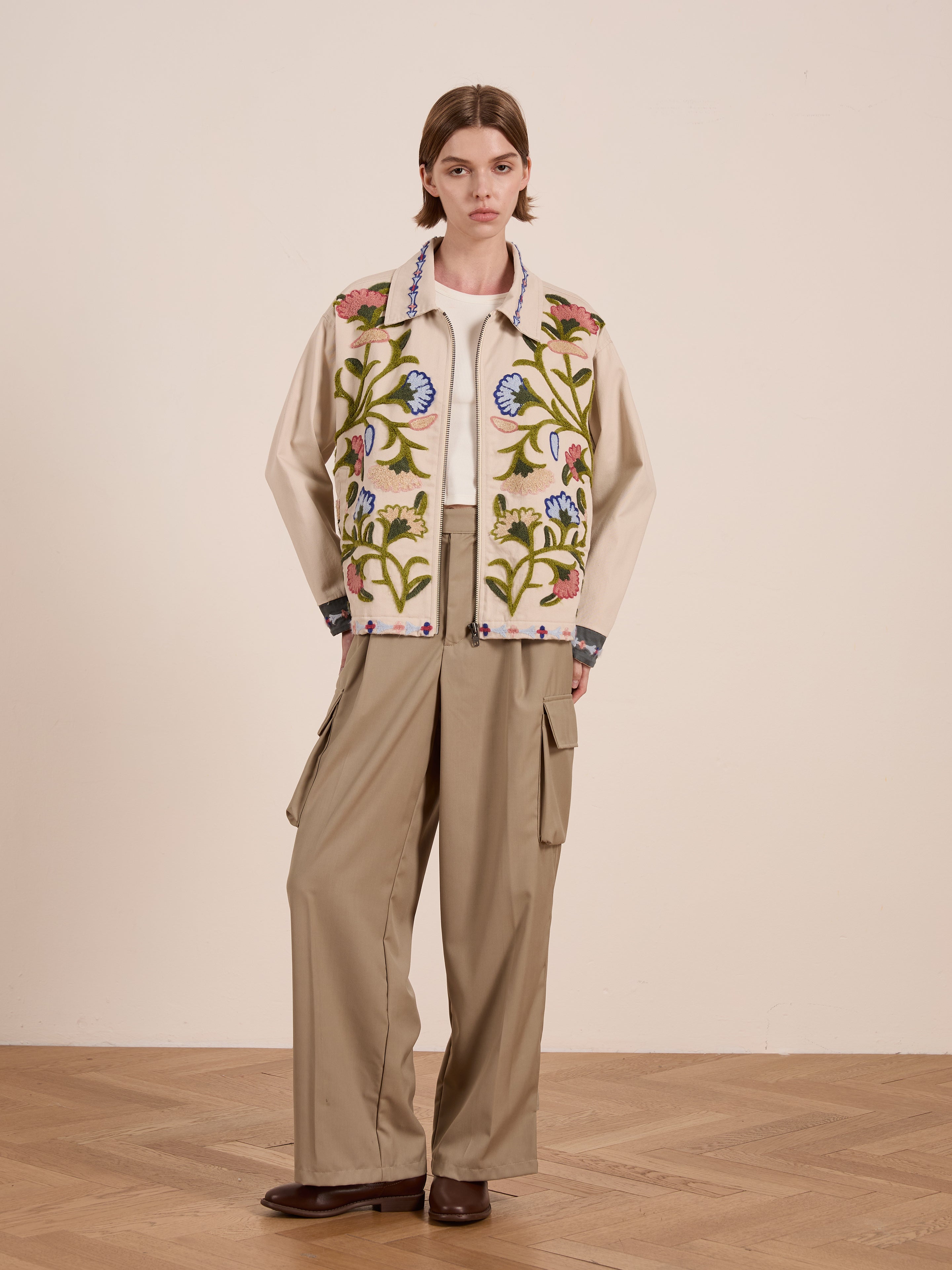 A person dressed in FOUND's Floral Embroidered Work Jacket over a beige shirt, paired with wide-leg khaki pants and brown shoes, stands on a wooden floor.