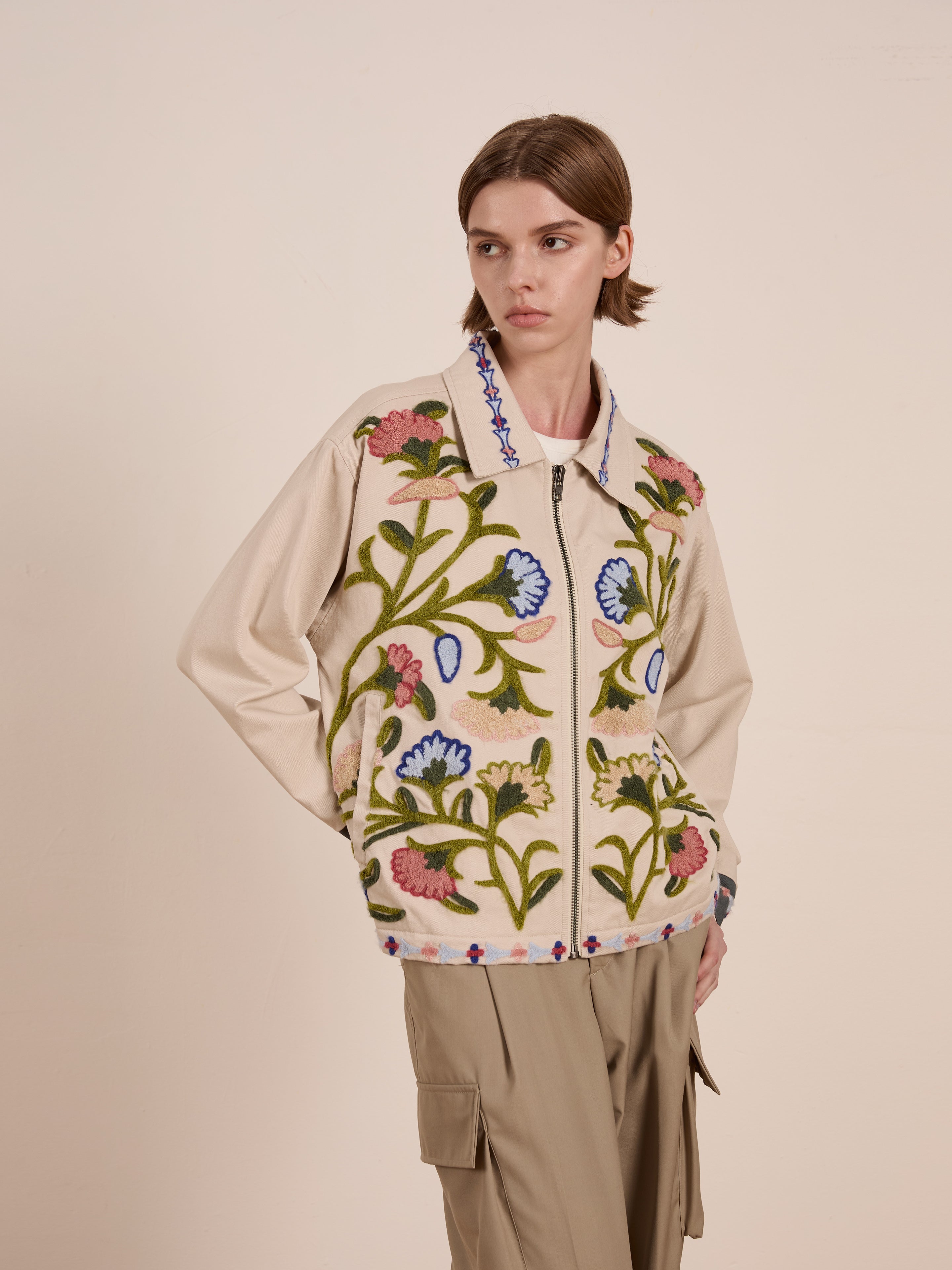 A person stands against a plain backdrop wearing FOUND's Floral Embroidered Work Jacket and beige pants, showcasing a chic blend of vibrant floral chainstitch embroidery with subtle utilitarian design elements.