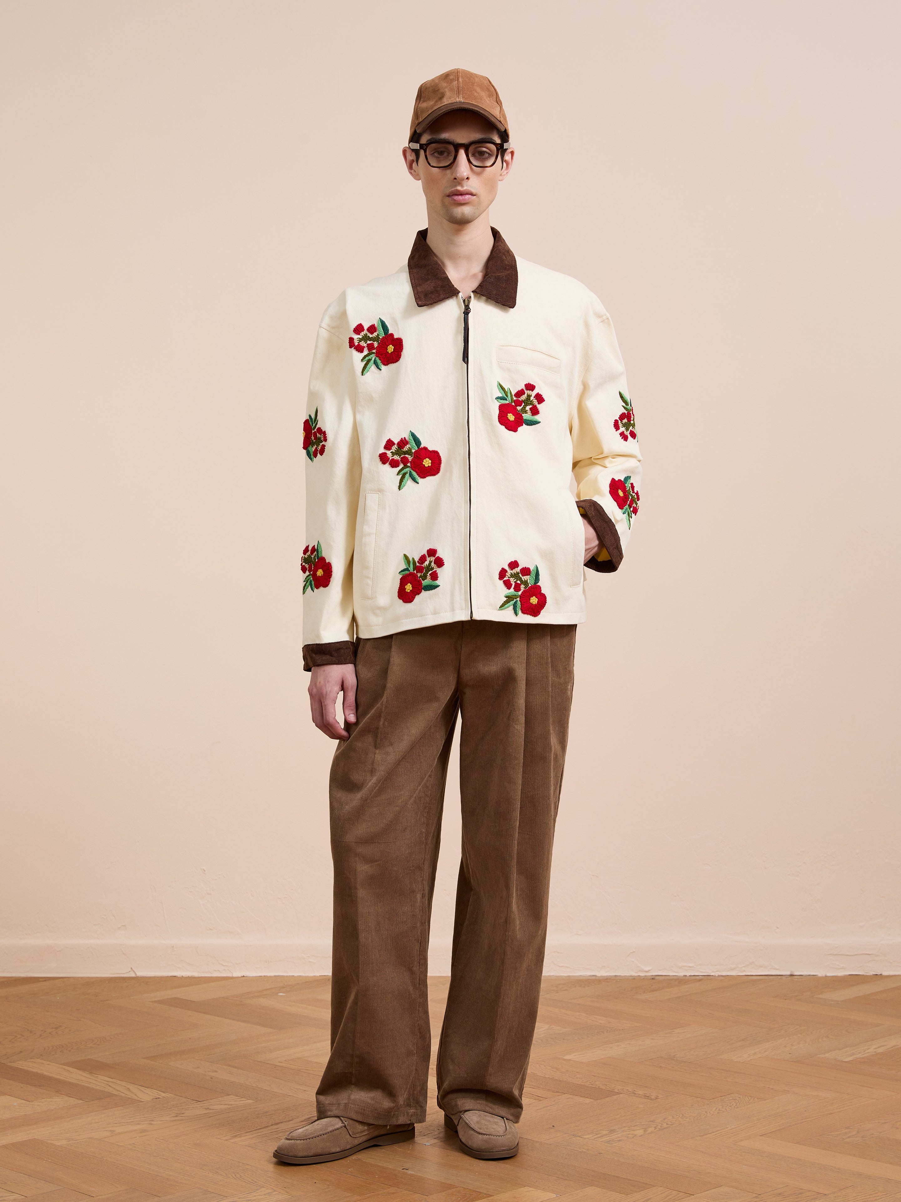 A person stands indoors against a beige background wearing Found's Floral Hand Embroidered Work Jacket with brown corduroy pants, brown shoes, a brown cap, and glasses.