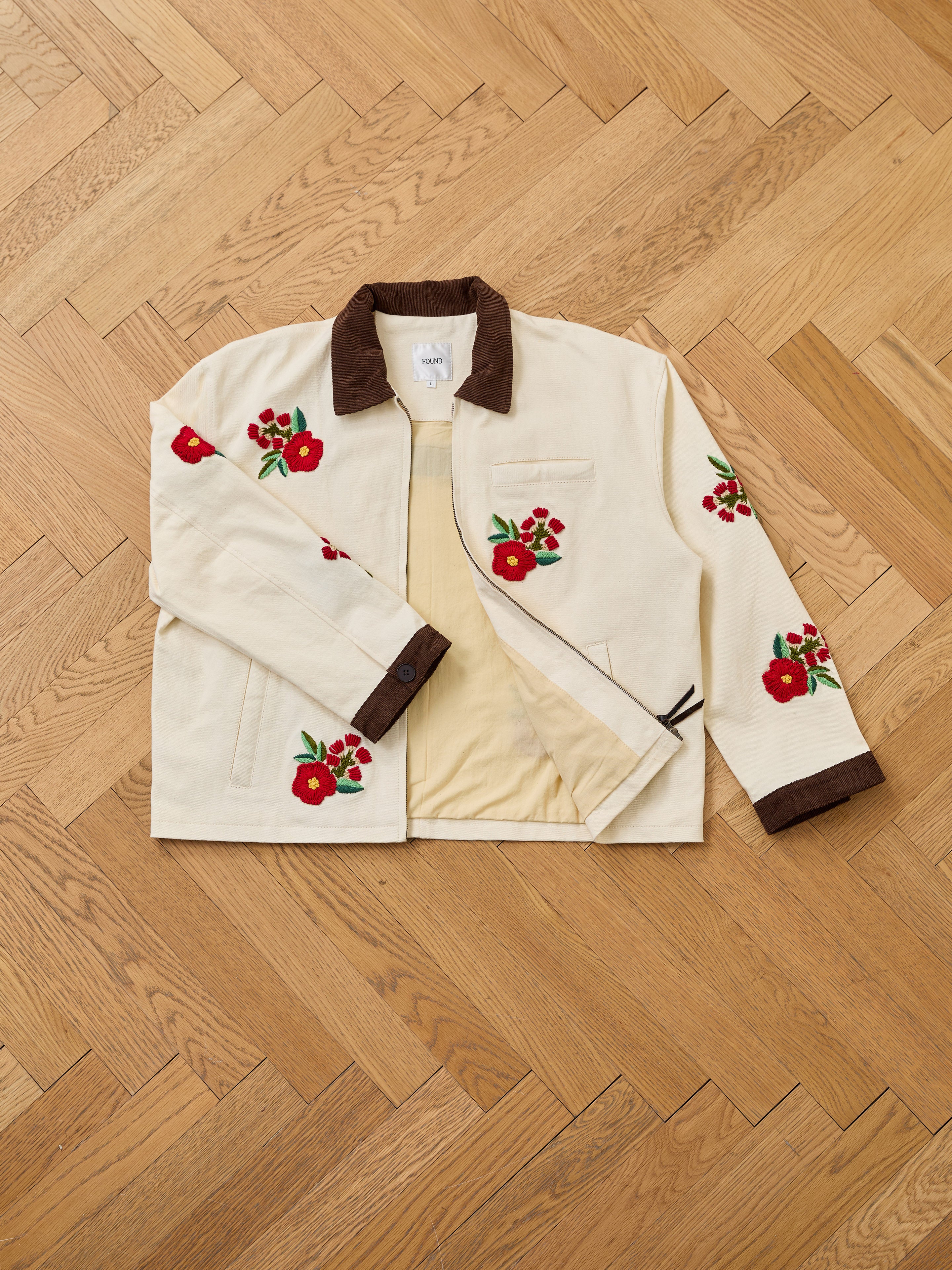 The Found Floral Hand Embroidered Work Jacket, in cream with brown collar and cuffs, vintage South Asian floral embroidery, and two front pockets, is shown laid flat on a wooden herringbone floor.
