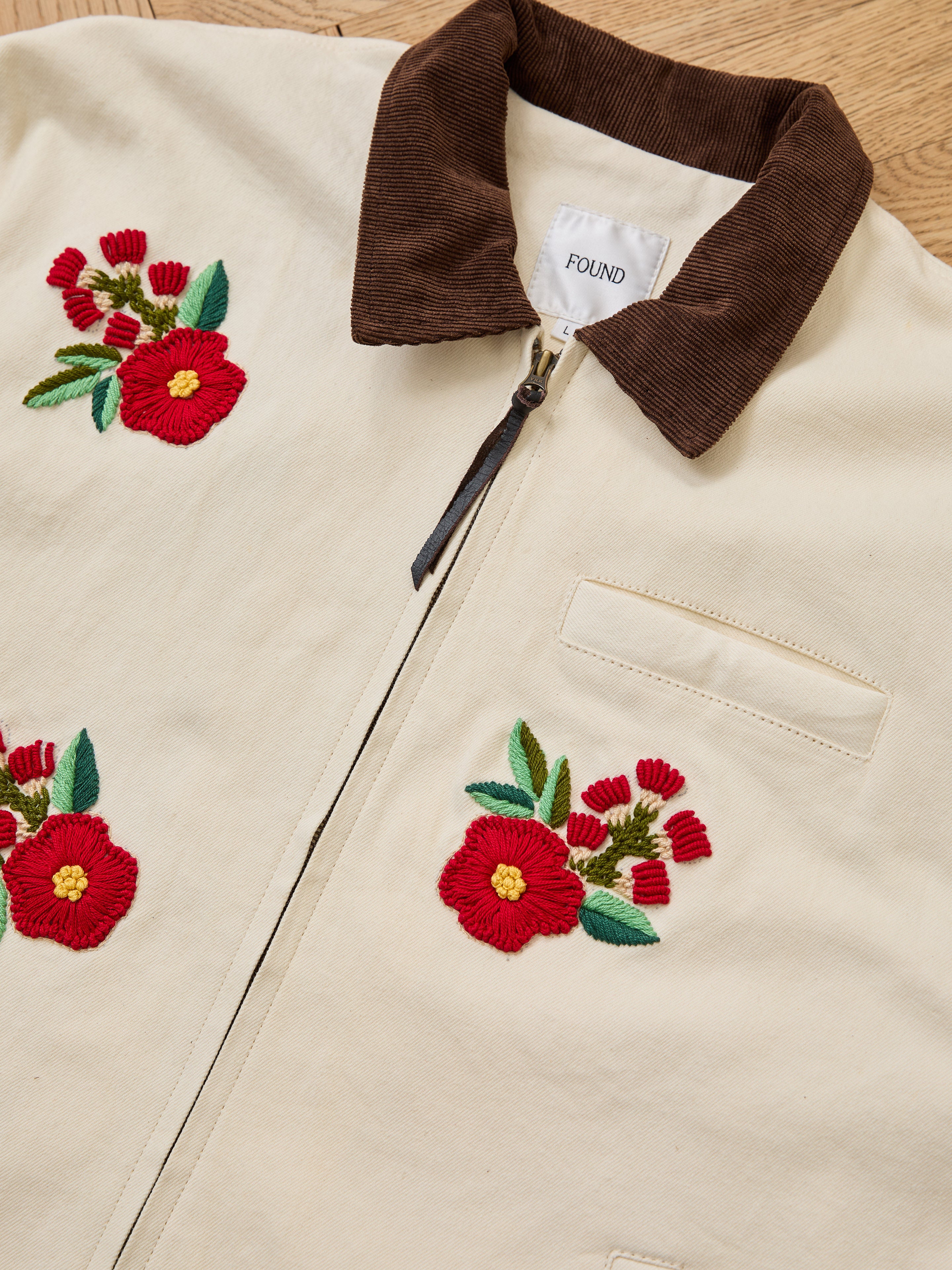 The Found Floral Hand Embroidered Work Jacket features cream fabric, brown corduroy collar, red South Asian embroidery, chest pocket, and brand label. Handcrafted in Pakistan and displayed flat on a wooden surface.
