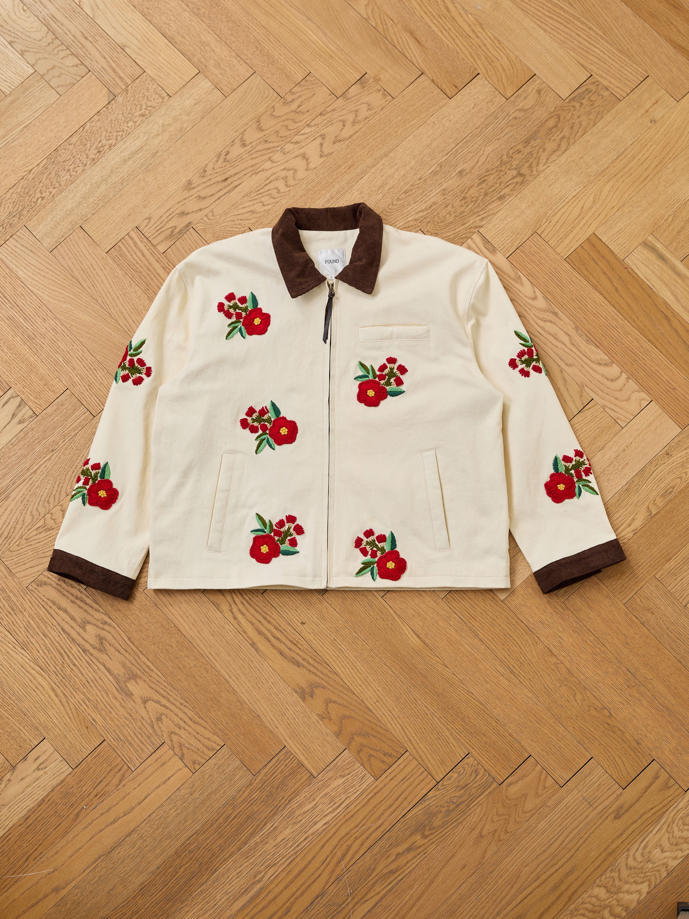 The Floral Hand Embroidered Work Jacket by Found, in cream with a brown collar and cuffs, showcases vintage South Asian needlework and red floral embroidery, displayed flat on a wooden herringbone floor.