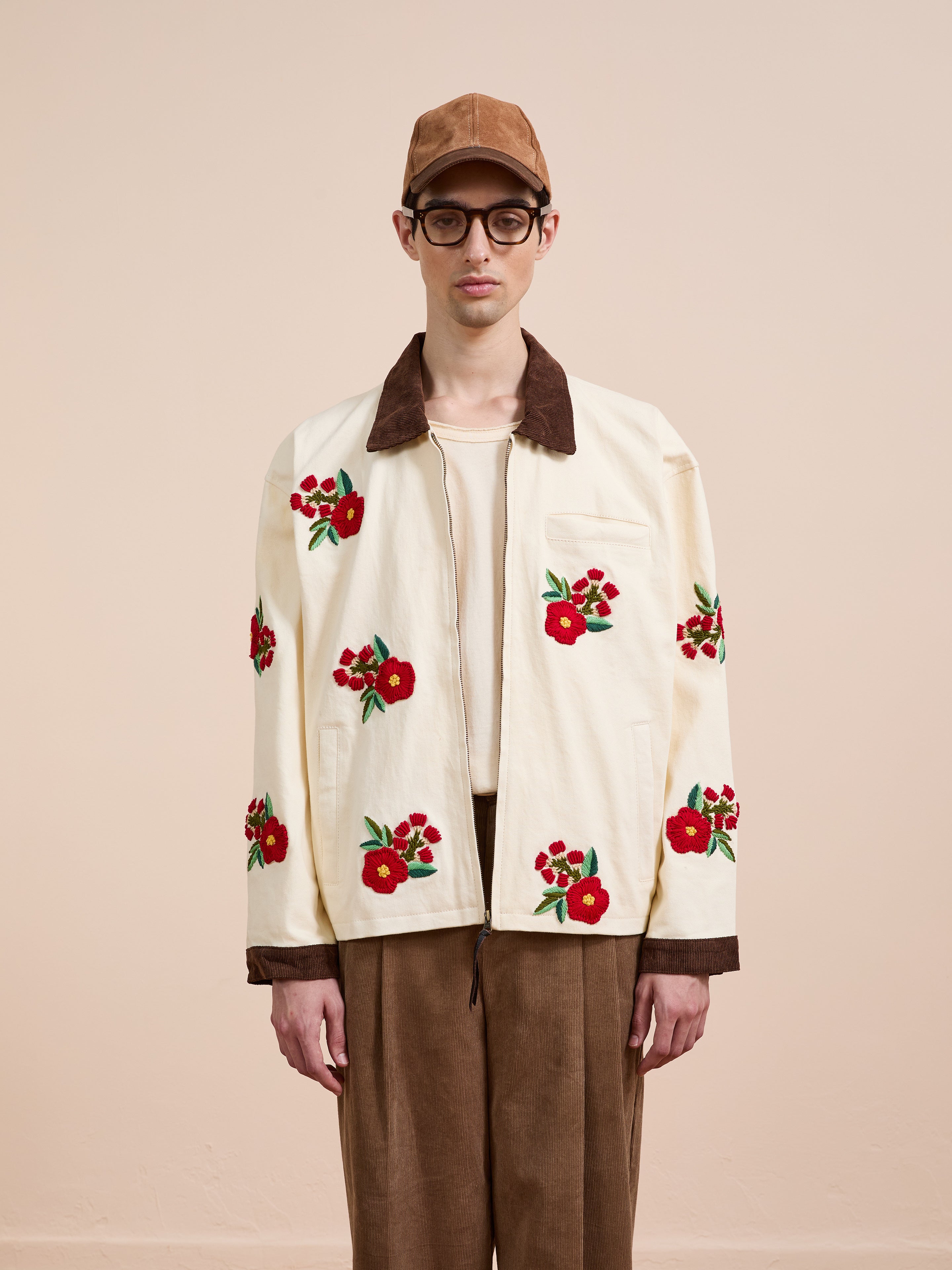 A person wearing a tan cap, glasses, brown pants, and the Found Floral Hand Embroidered Work Jacket stands against a plain beige background.