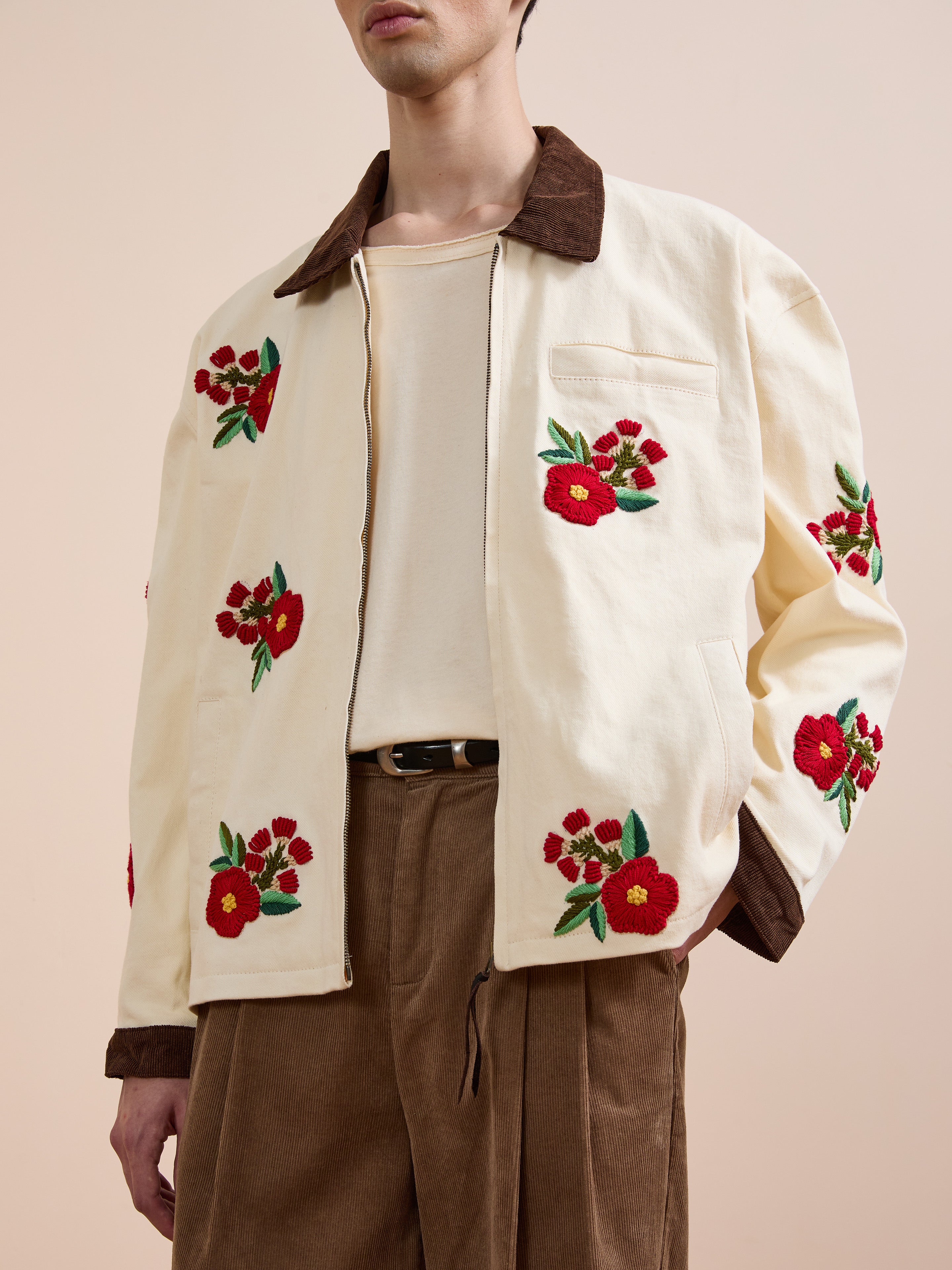 A person models the Found Floral Hand Embroidered Work Jacket with red flower motifs, paired with a cream shirt and brown trousers, against a beige background—highlighting vintage South Asian craftsmanship.