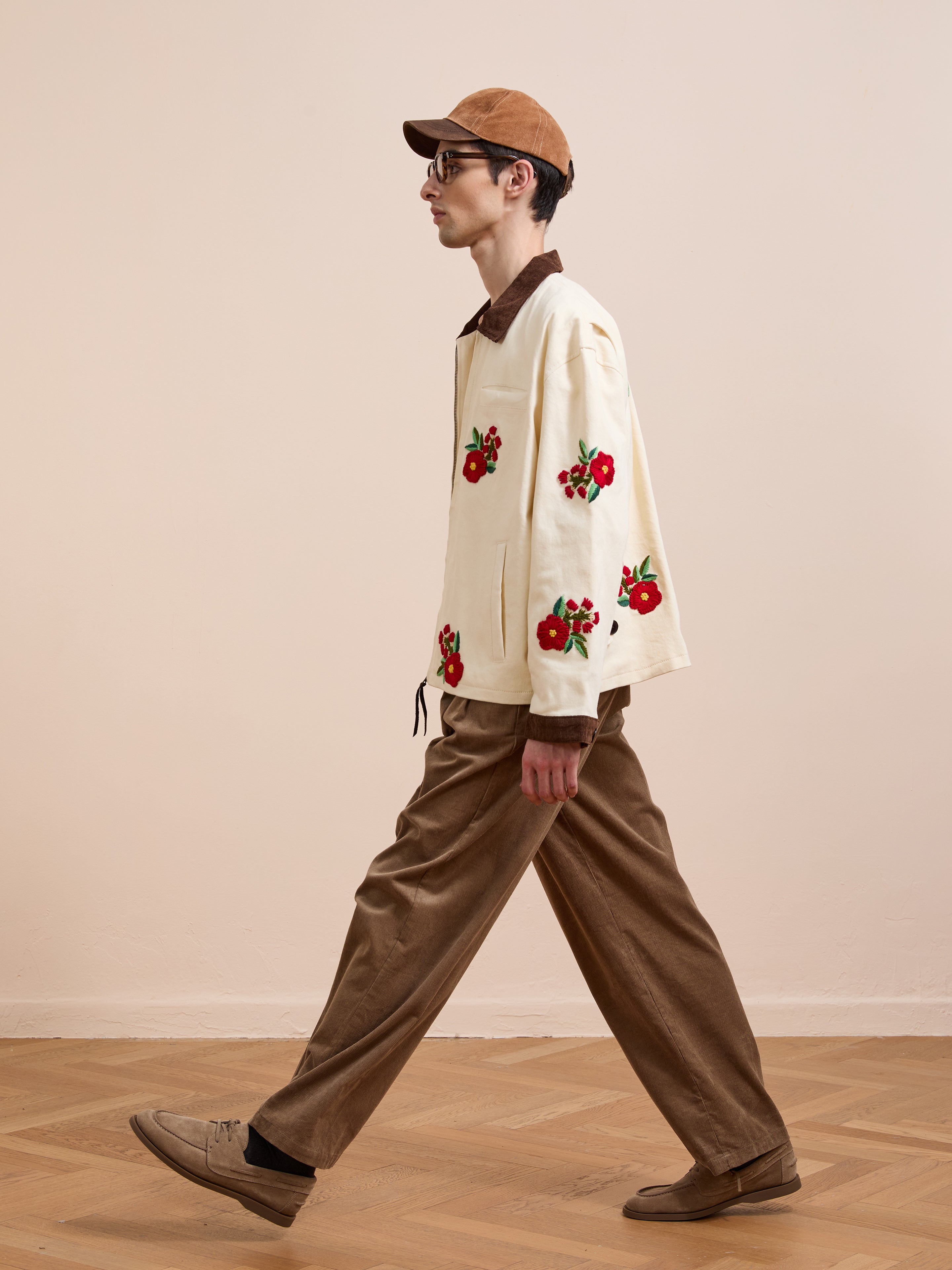 A person wears the Found Floral Hand Embroidered Work Jacket, styled with brown corduroy pants, brown shoes, and a brown cap while walking indoors on a wooden floor.