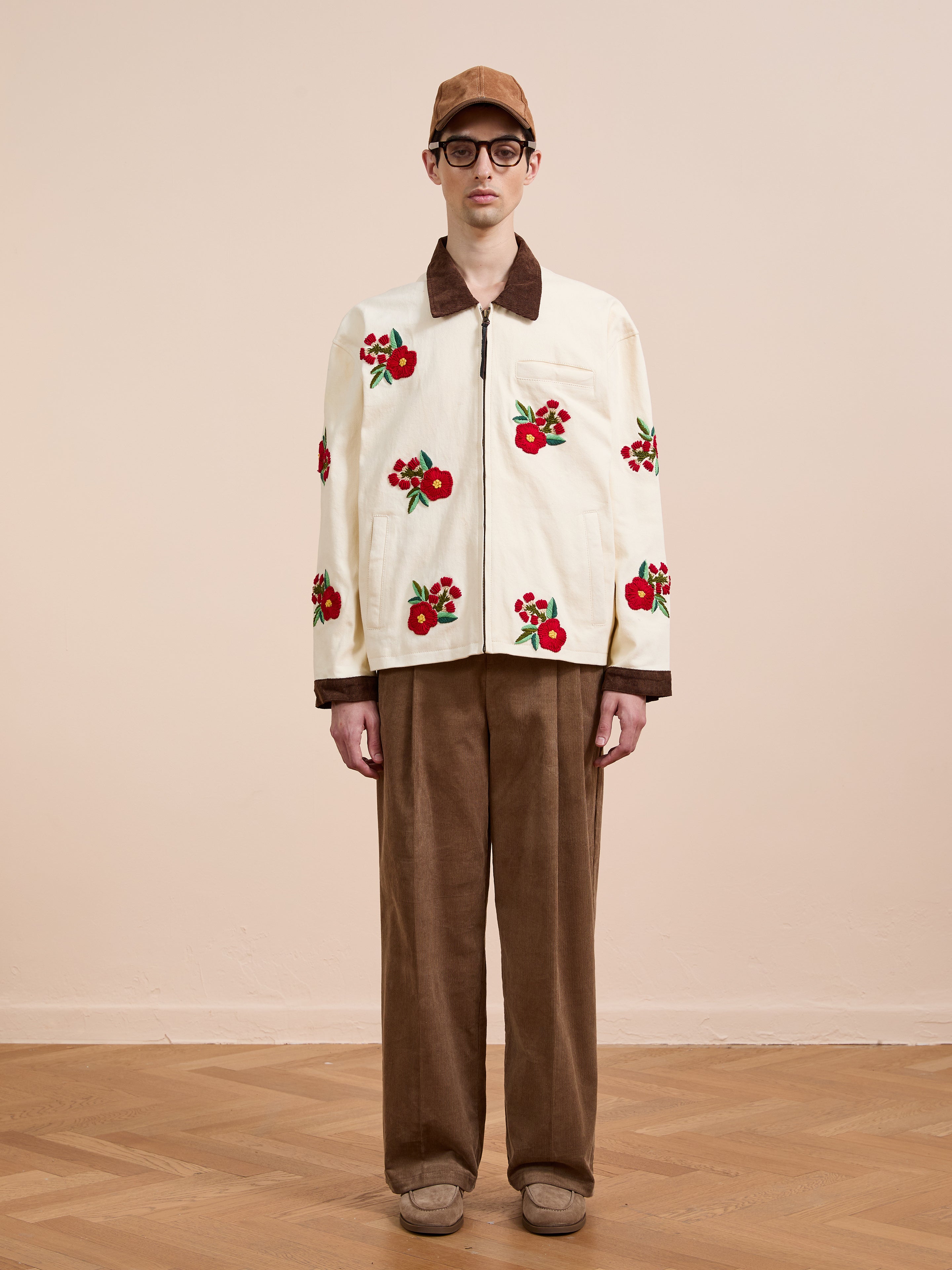 A man stands indoors against a neutral background, wearing Found’s Floral Hand Embroidered Work Jacket with vintage South Asian-inspired cream floral embroidery, brown collar and cuffs, brown corduroy pants, cap, and glasses.