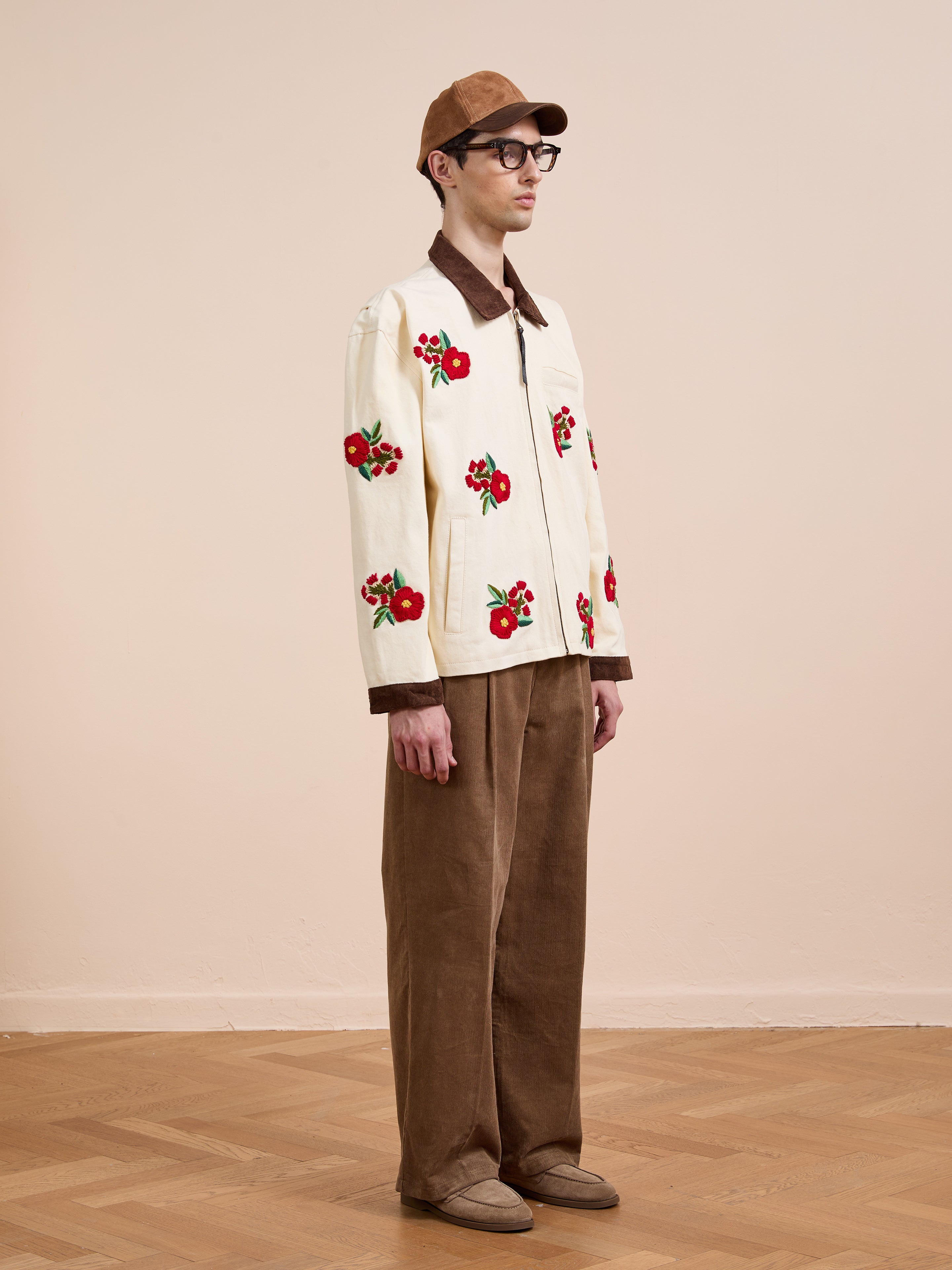 A man in glasses and a tan cap stands indoors on a wooden floor against a beige wall, wearing Found's Floral Hand Embroidered Work Jacket inspired by vintage South Asian needlework.