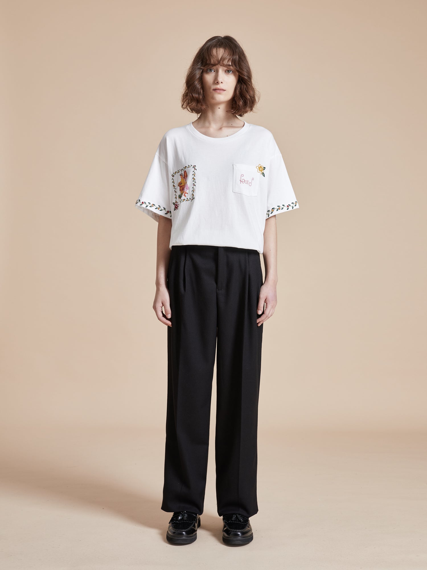 The model is wearing a white Flower Children Tee with floral Phulkari style embroideries and black trousers.