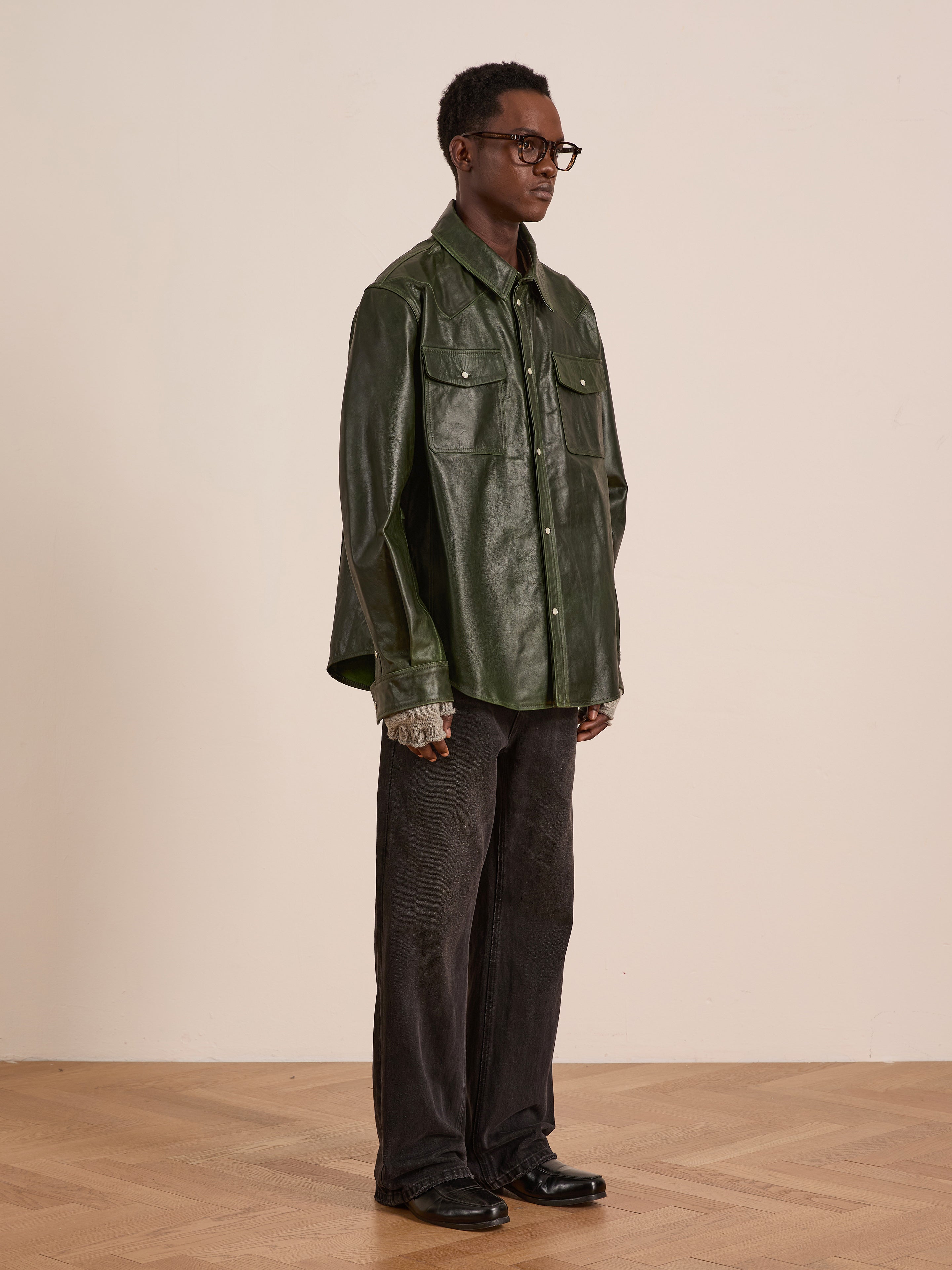 Wearing the FOUND Forest Leather Western Shirt, black wide-leg pants, glasses, and black shoes, a person stands indoors on a wooden floor against a plain light-colored wall.