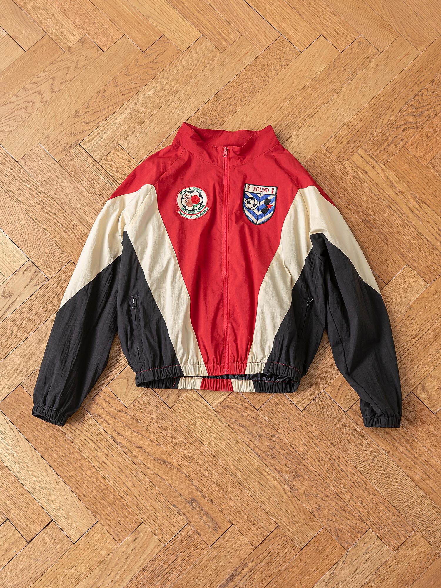 The Pine and Rose Track Jacket by FOUND, in red, beige, and black with two embroidered patches (a round text patch and a shield), offers unisex sportswear style and versatility. Displayed on a wooden floor.