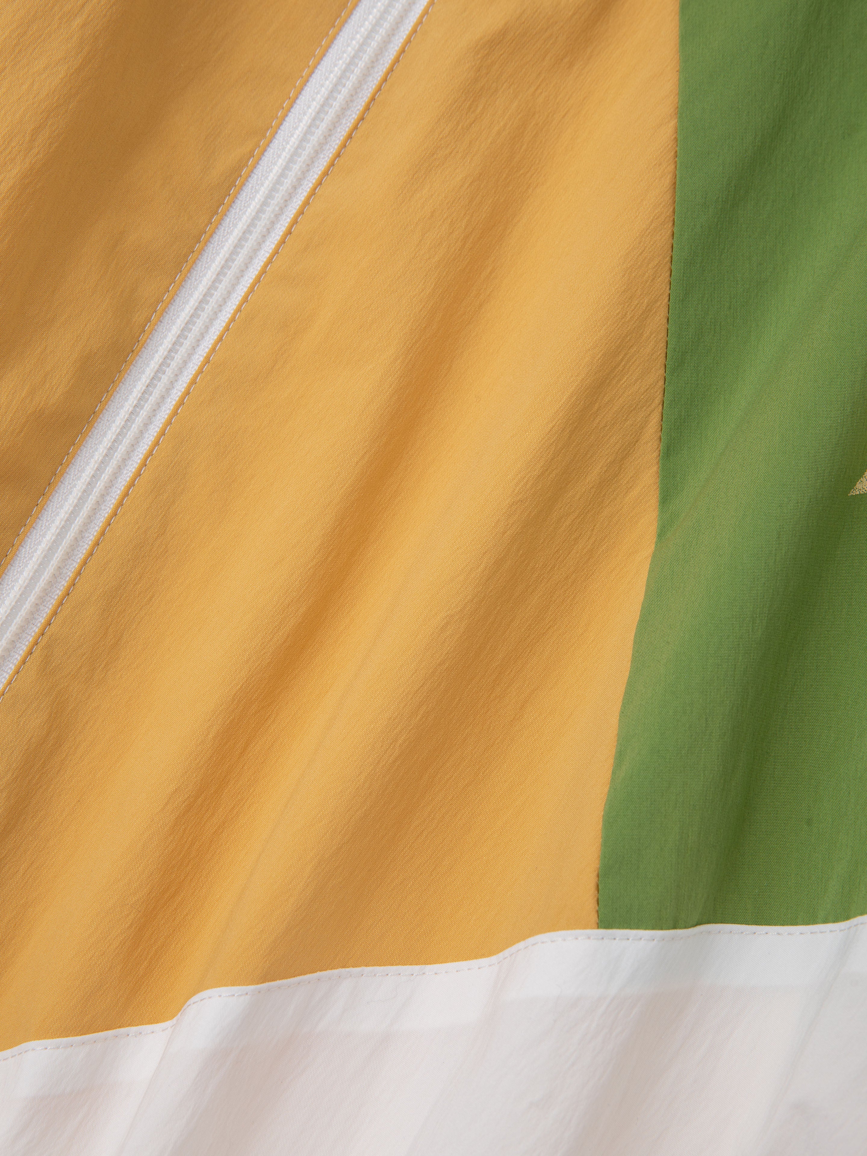Close-up of fabric from FOUND's Legacy Cricket Track Jacket, showcasing yellow, green, and white sections with a diagonal white zipper—reminiscent of vintage athletic gear.