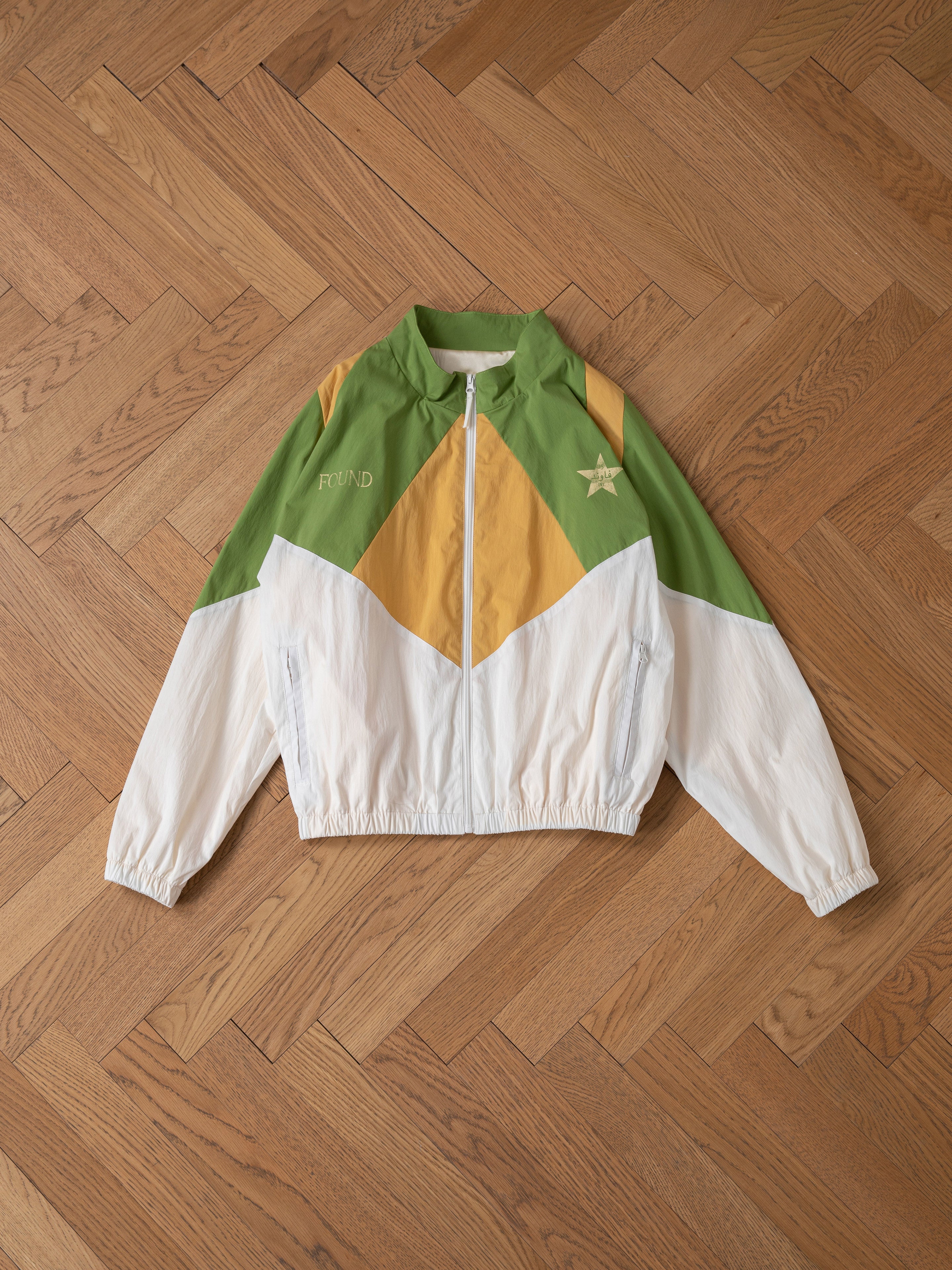 The Legacy Cricket Track Jacket by FOUND features green, yellow, and white sections with a star and "ROUND" text, reminiscent of retro international tracksuits, stylishly displayed on a parquet wooden floor.