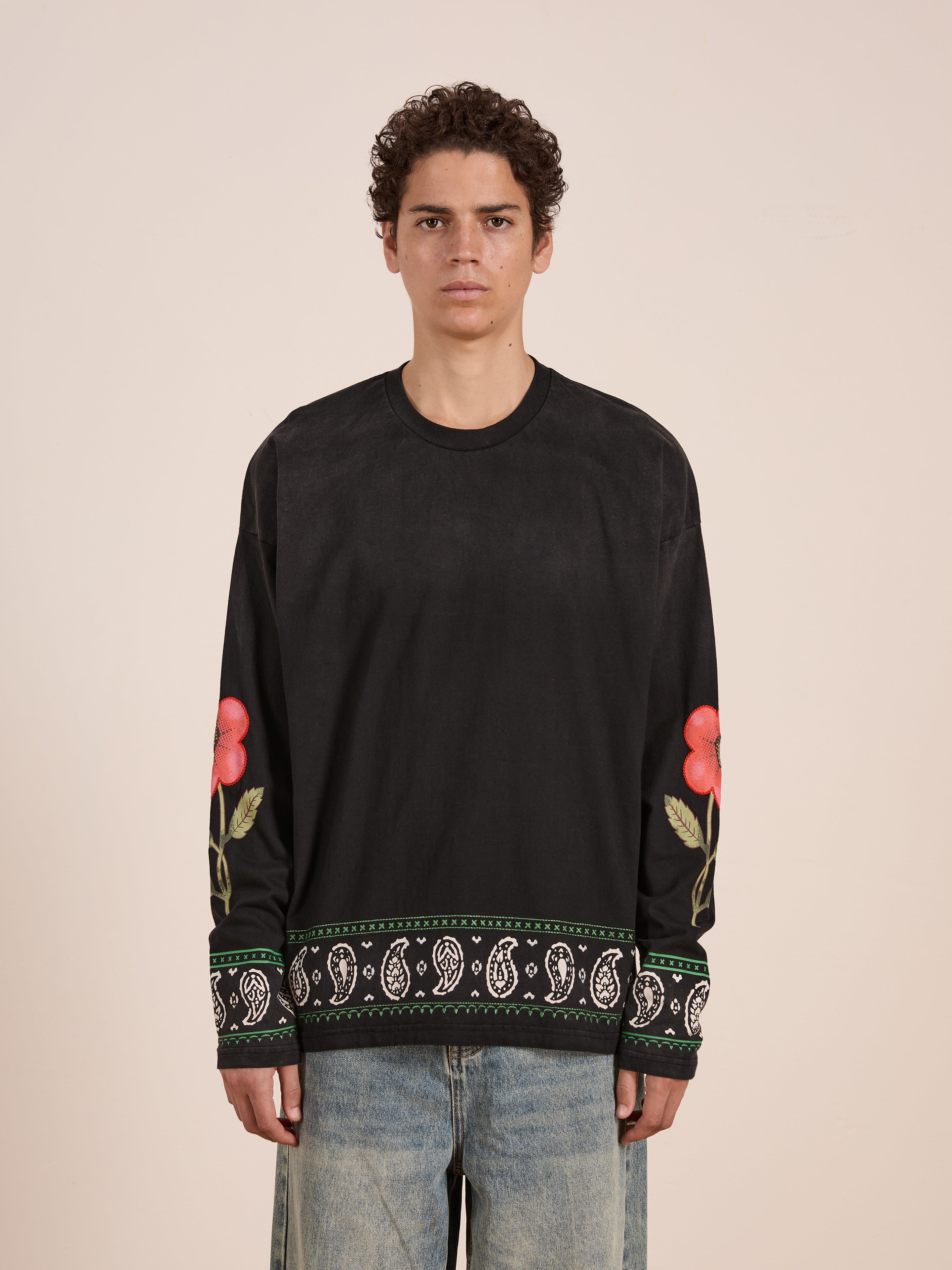 A person wears the FOUND Garden Bloom Embroidered LS Tee, a black long-sleeve with floral embroidery and paisley trim at the cuffs and hem, paired with light blue jeans, standing against a plain background in a relaxed fit.