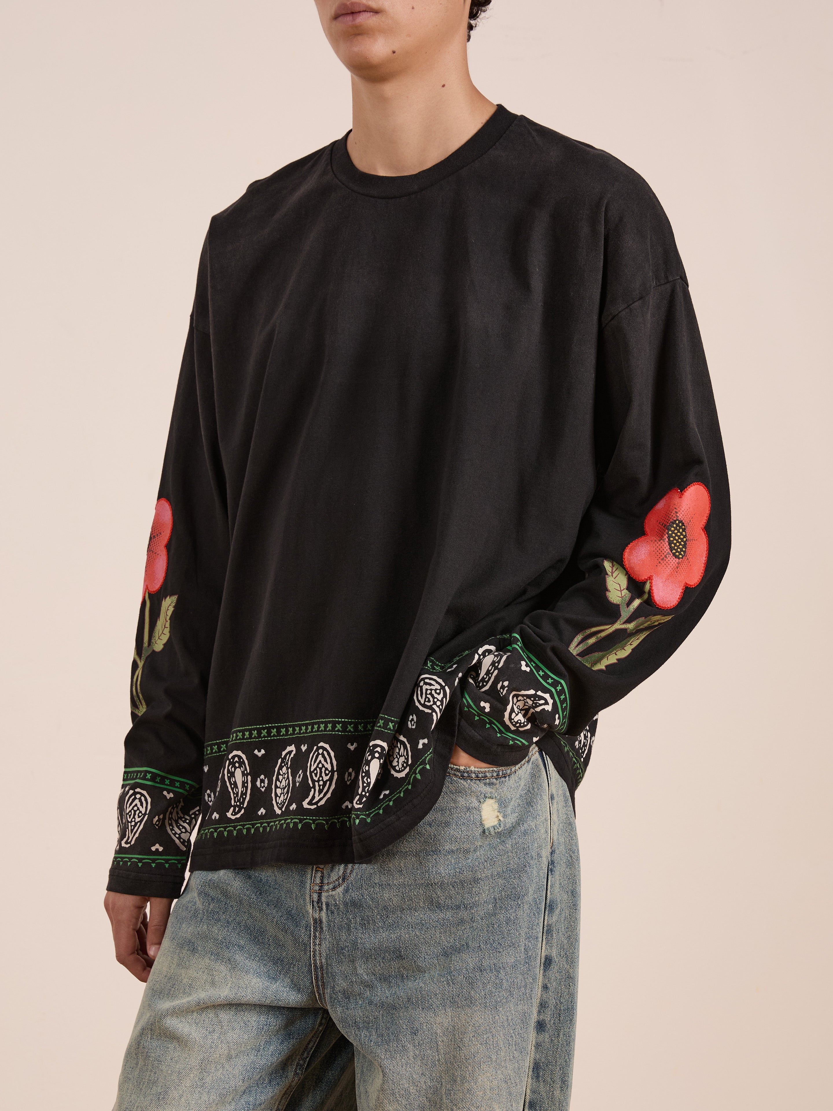A person wears the FOUND Garden Bloom Embroidered LS Tee, featuring a relaxed fit, black fabric with red floral embroidery and bandana-inspired details at the cuffs and hem, paired effortlessly with light blue jeans.