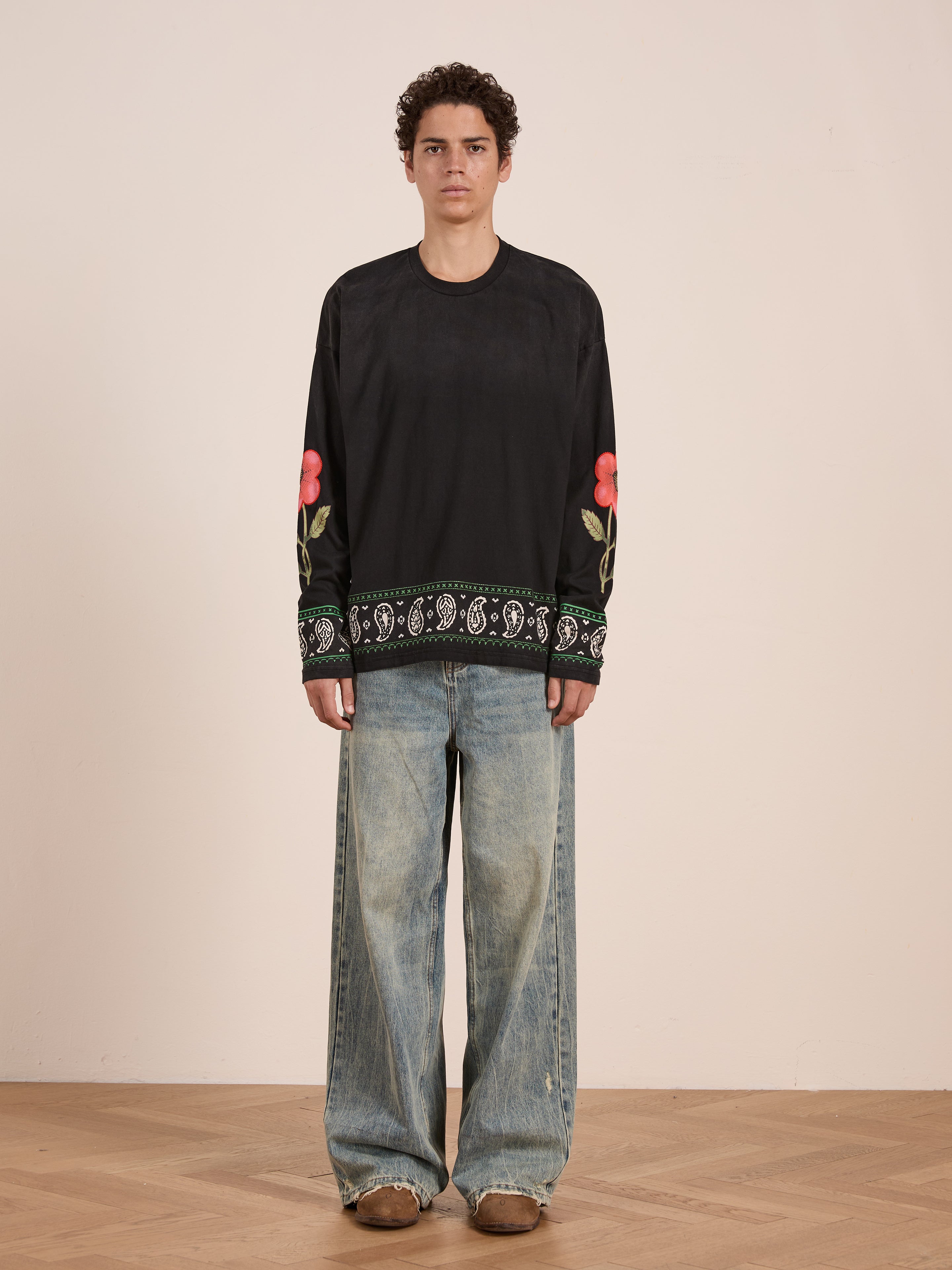 A person stands indoors facing the camera in a neutral expression, wearing the FOUND Garden Bloom Embroidered LS Tee with floral sleeve embroidery and a relaxed fit, paired with wide-leg blue jeans.