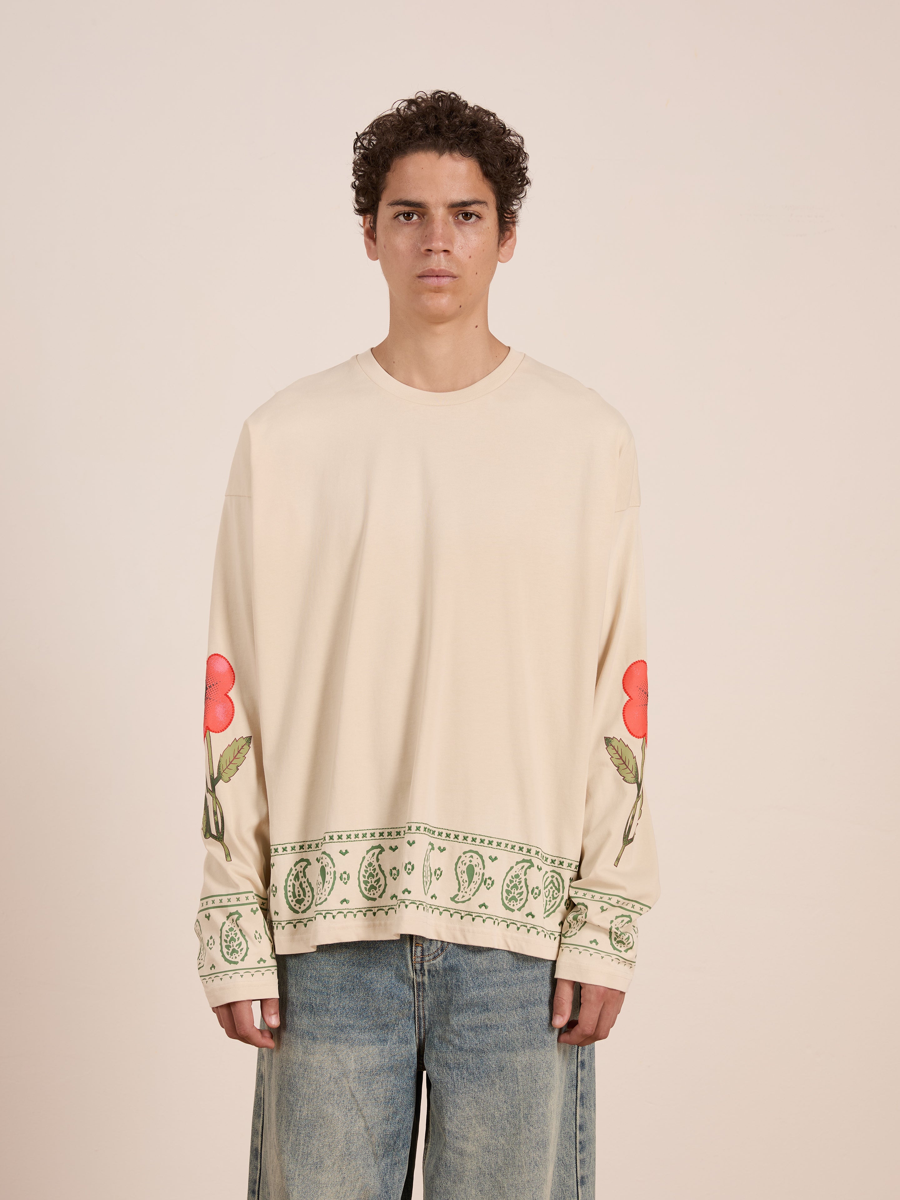 A young person wears the FOUND Garden Bloom Embroidered LS Tee, a unisex relaxed fit with embroidered red flowers on the sleeves and green paisley details at the cuffs and hem, paired with light blue jeans.
