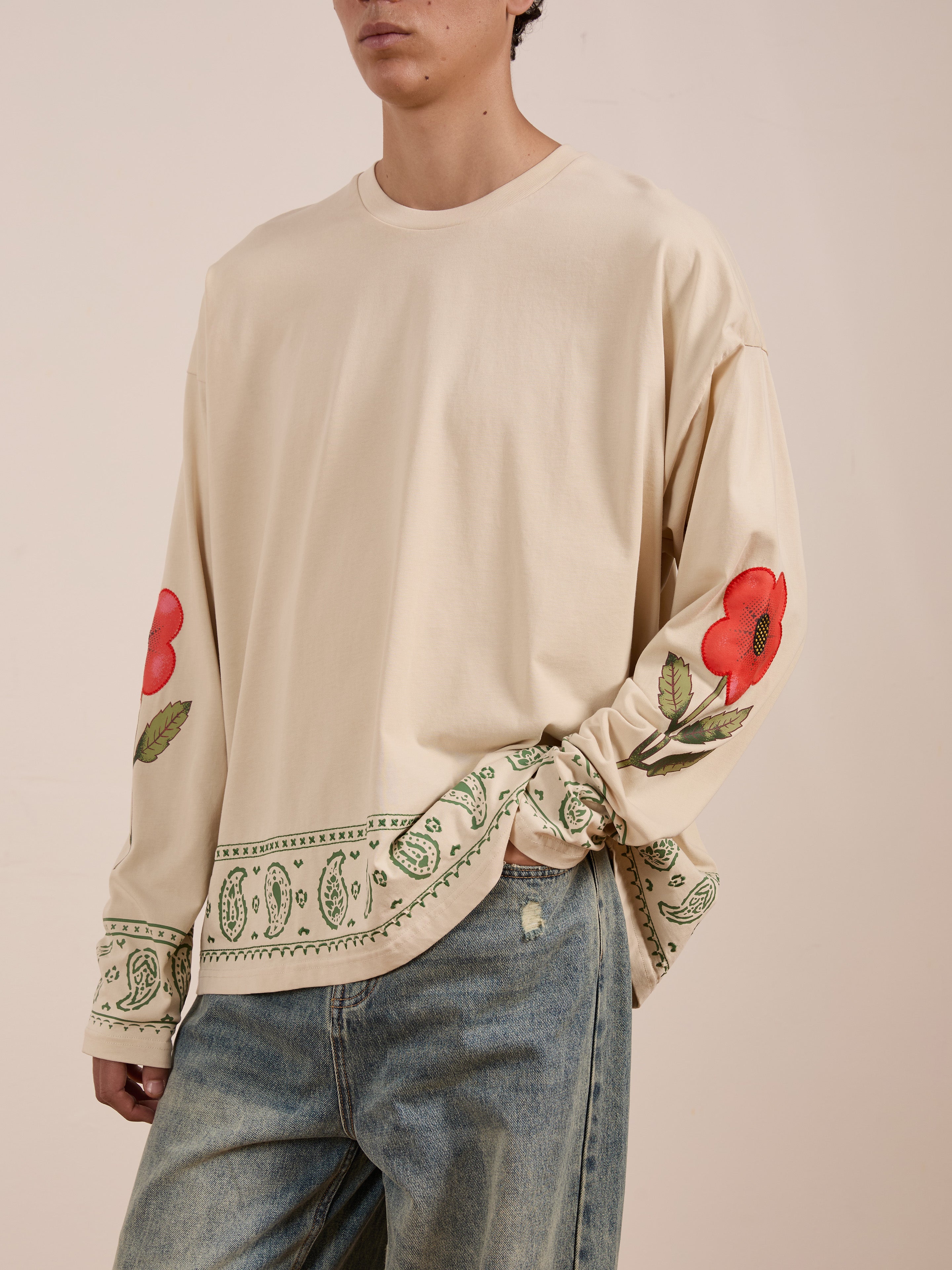 Person wearing the FOUND Garden Bloom Embroidered LS Tee, a unisex relaxed-fit beige long-sleeve with red and green floral embroidery on sleeves and hem, paired with blue jeans.