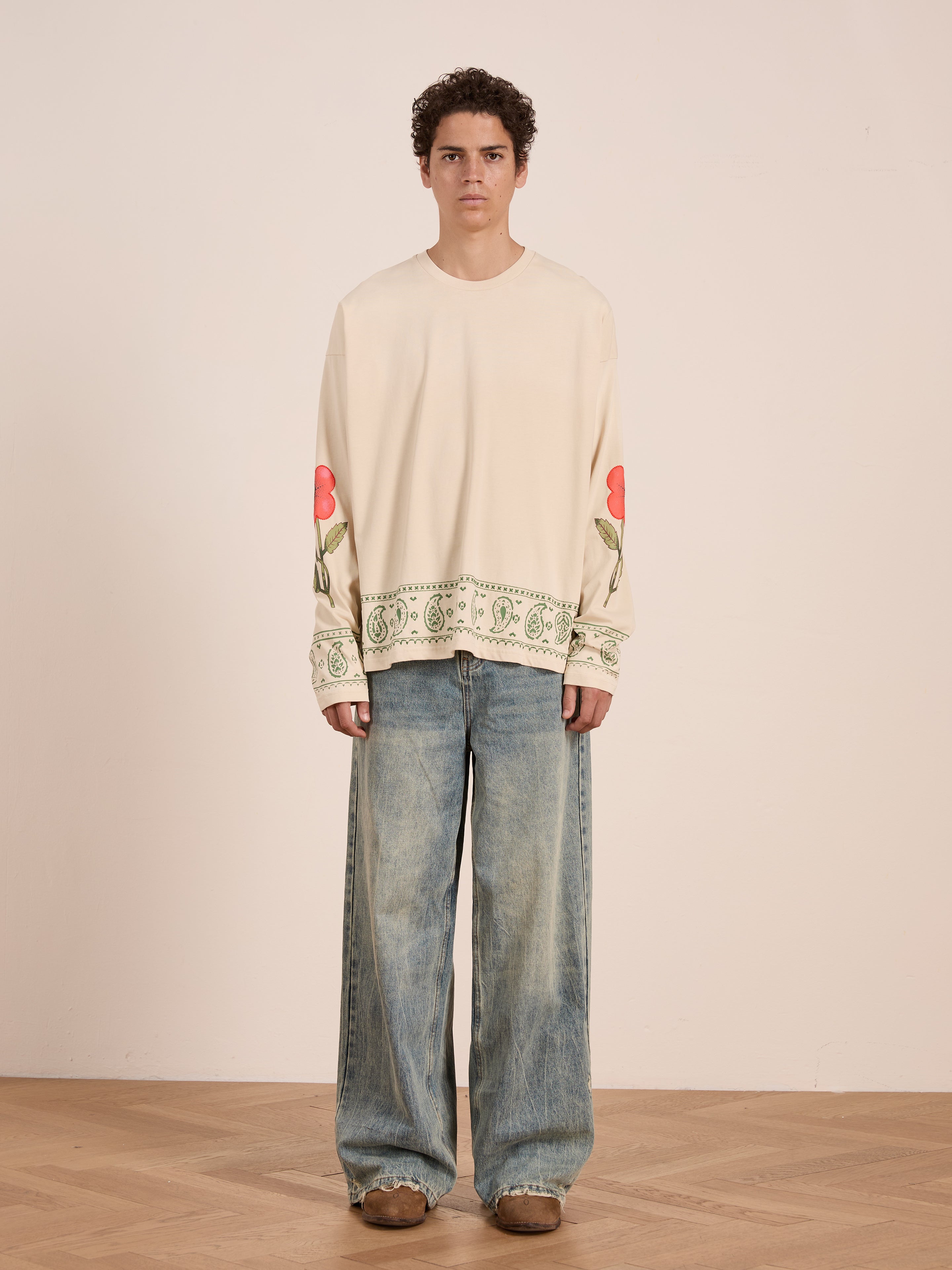 Indoors, a person wears the FOUND Garden Bloom Embroidered LS Tee in cream with floral embroidery, paired with wide-leg blue jeans and brown shoes. The unisex relaxed fit enhances the laid-back vibe against a plain wall.