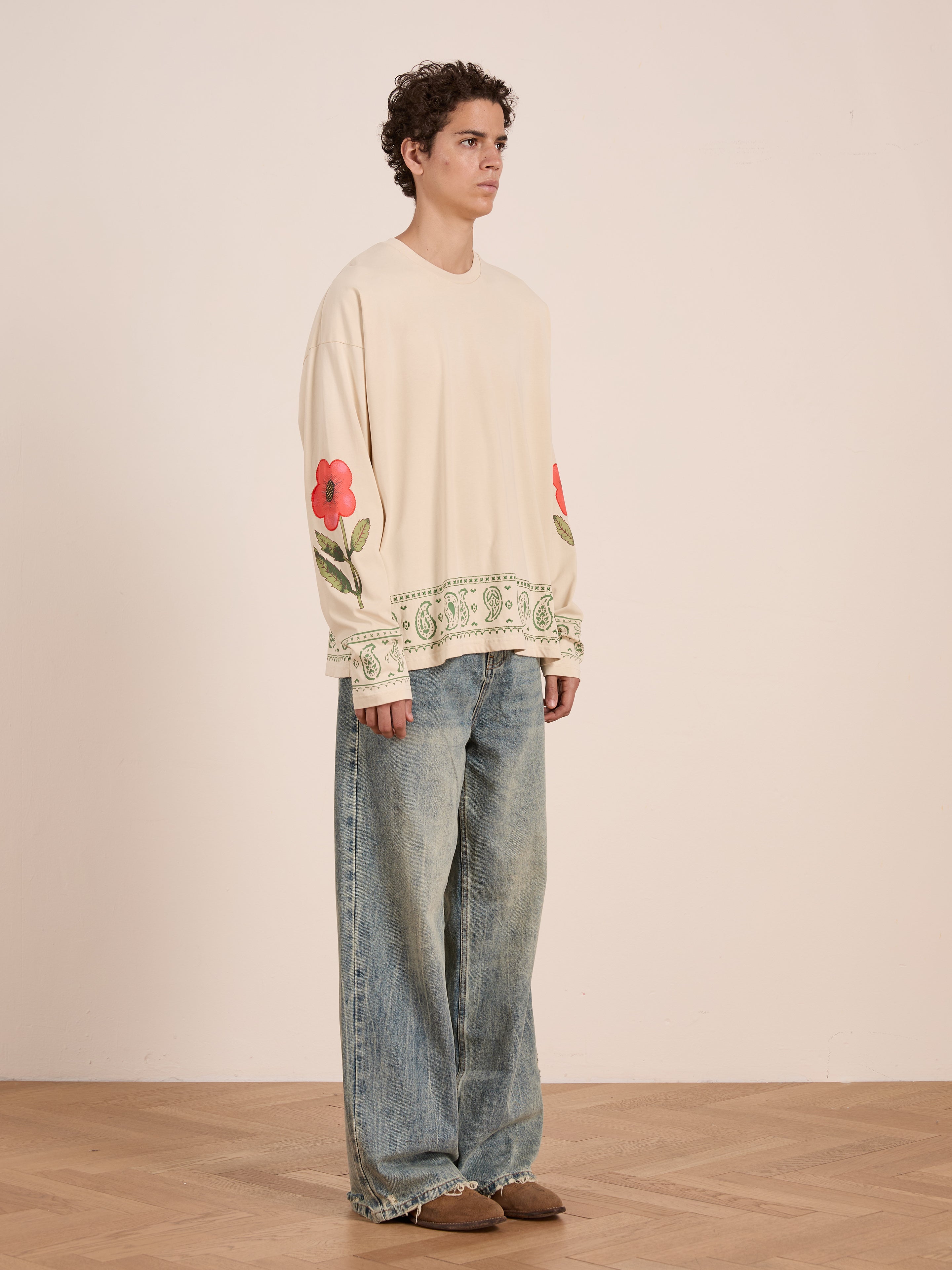 A person stands indoors in a cream FOUND Garden Bloom Embroidered LS Tee with floral embroidery and wide-leg blue jeans, looking slightly to the side in a relaxed unisex fit.