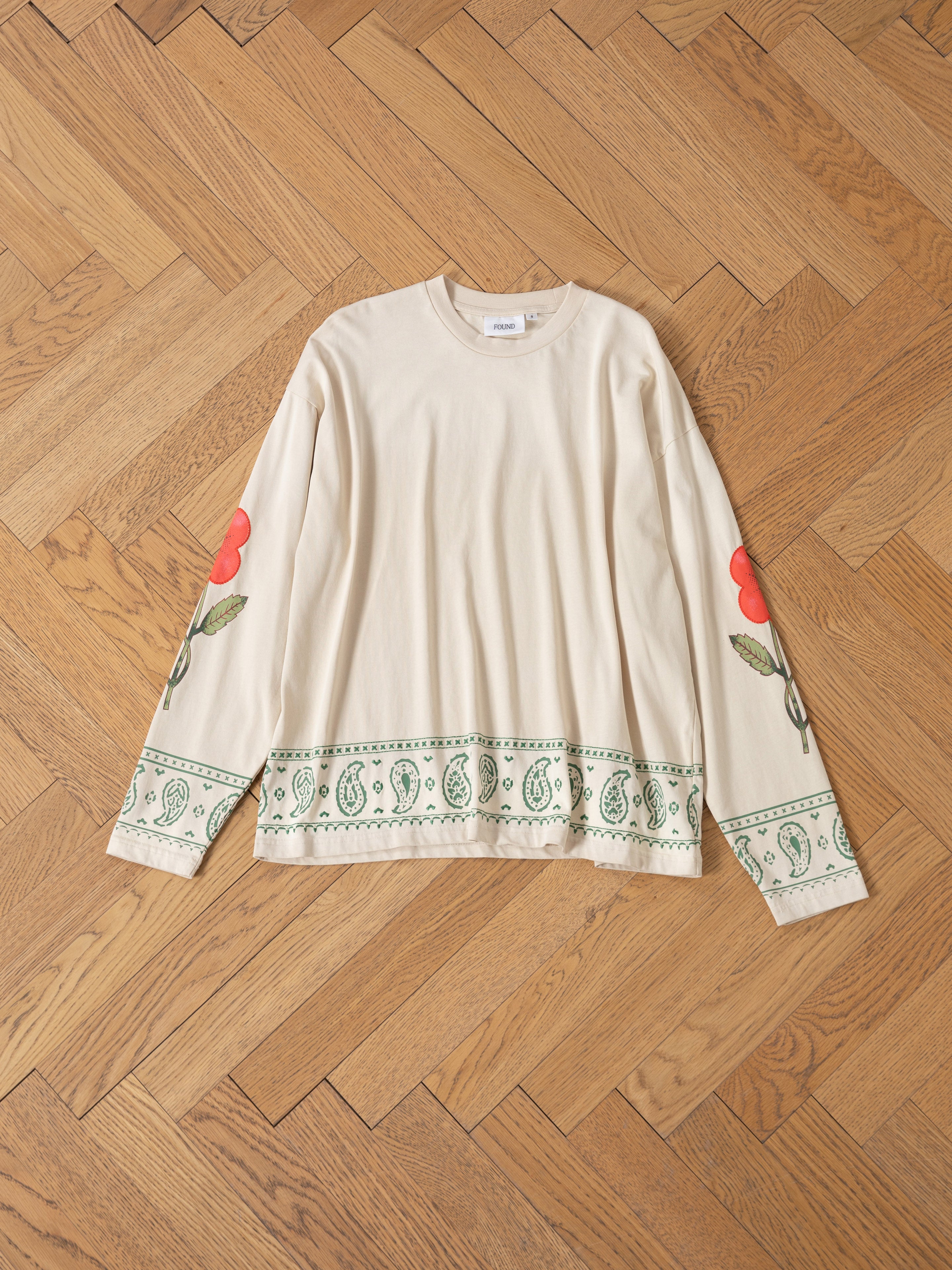 The FOUND Garden Bloom Embroidered LS Tee has a unisex relaxed fit, with red flower and green leaf embroidery on the sleeves and a green paisley border at the hem, shown laid flat on a wooden herringbone floor.
