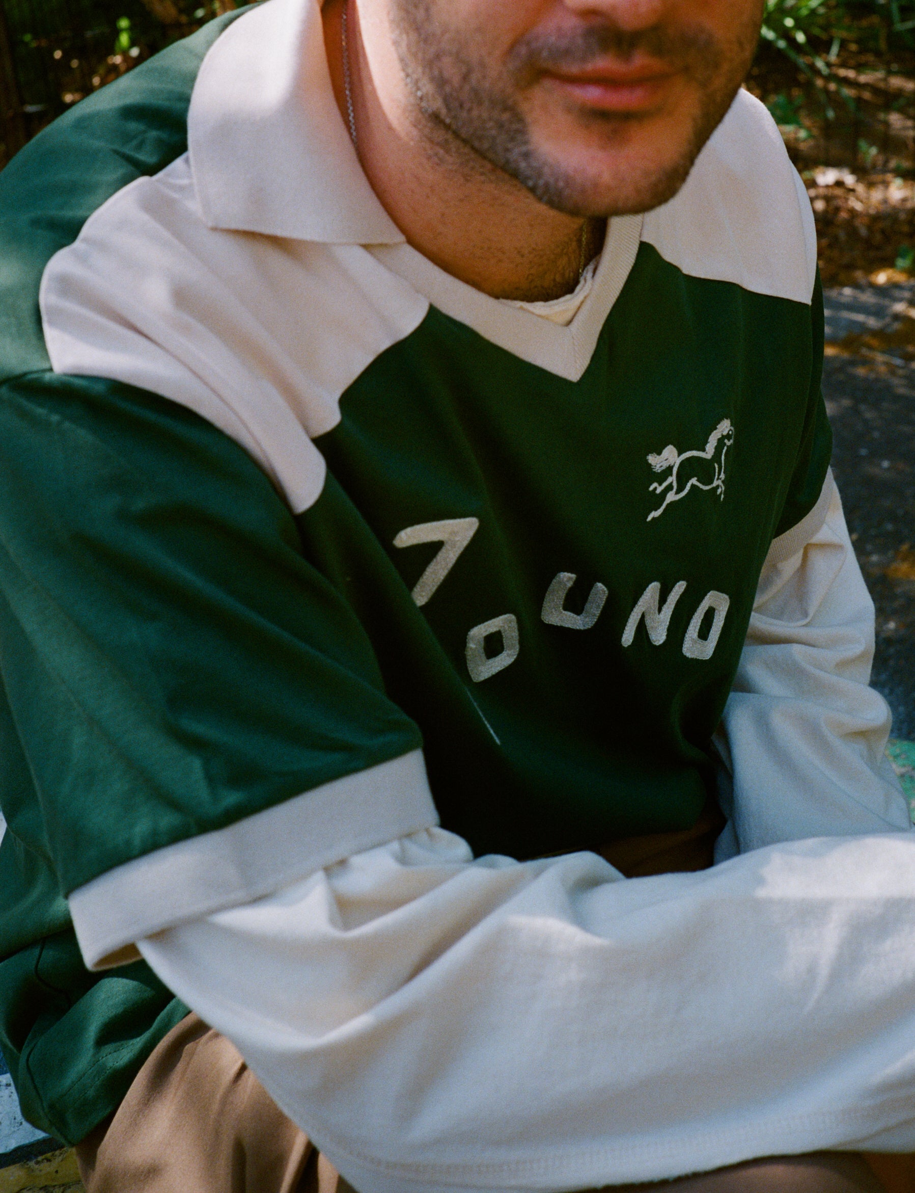 A man sits outdoors wearing the FOUND Goji Green Jersey, a green and white collared shirt inspired by vintage athletic uniforms with "FOUND" and an animal graphic on it.