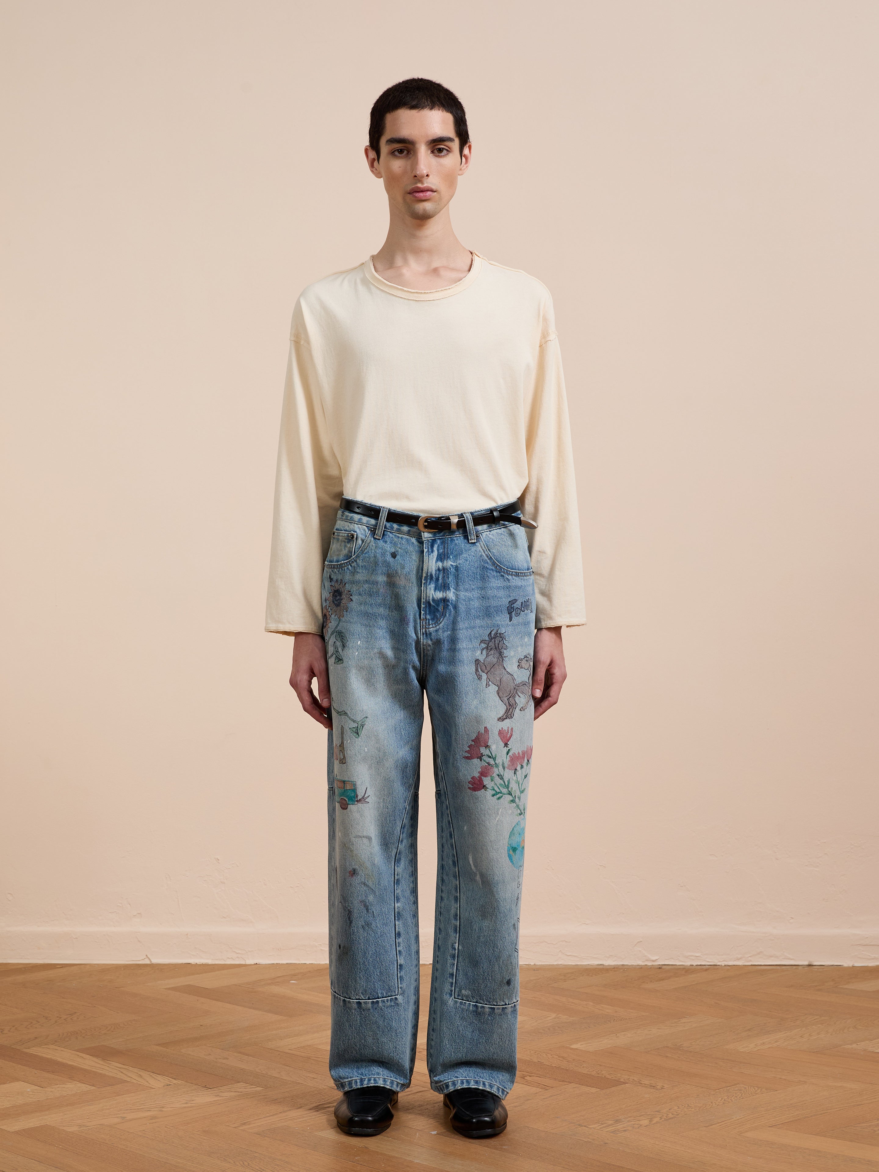 Indoors, a person wears FOUND Hand Art Jeans—loose, light blue denim with original artwork—paired with a long-sleeve cream shirt, black belt, and black shoes against a beige wall and wood floor.