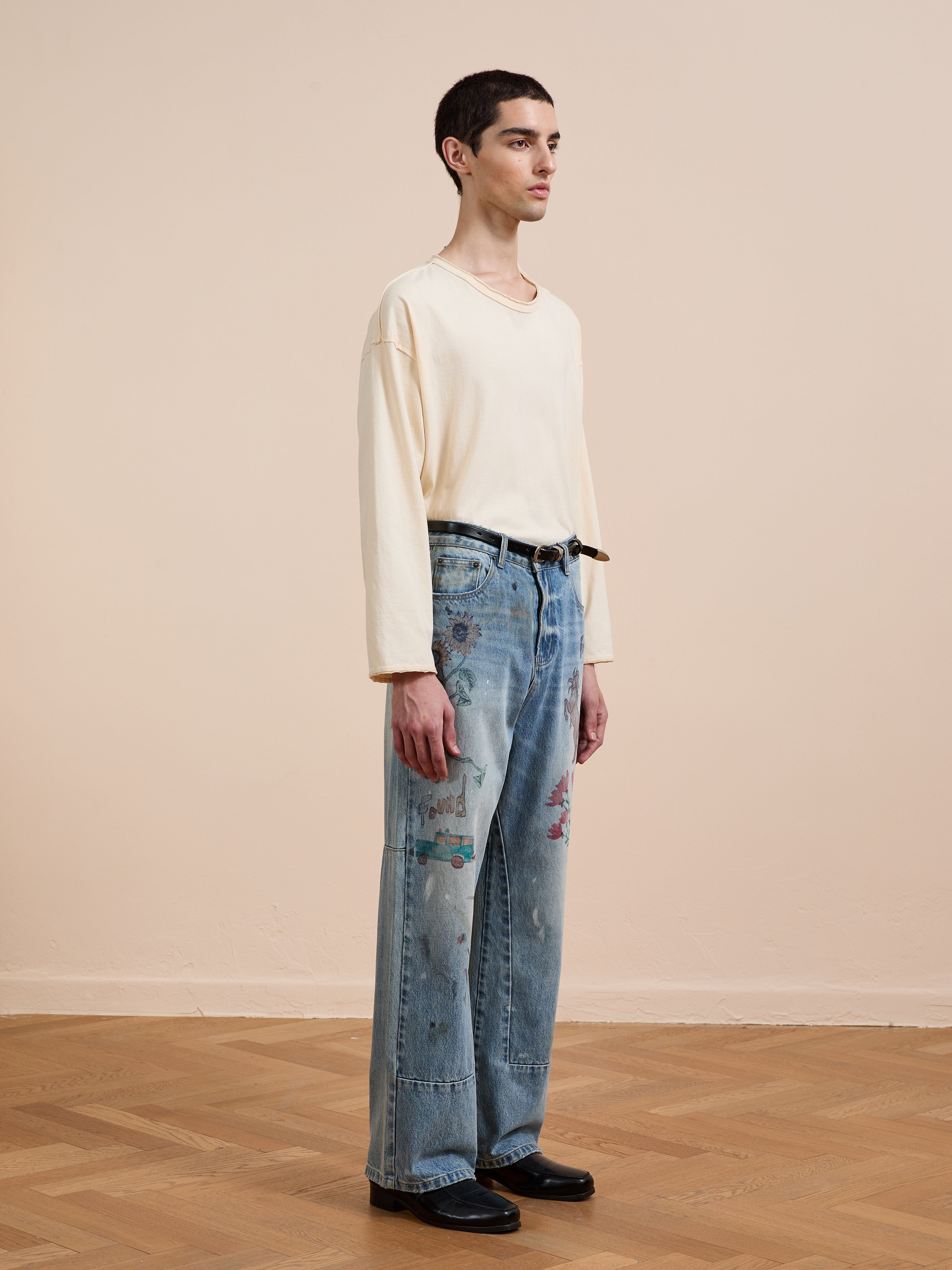 A person stands indoors on a wooden floor, facing left in profile, wearing FOUND’s Hand Art Jeans featuring blue denim with original hand-drawn artwork, paired with a cream long-sleeve shirt and black shoes.