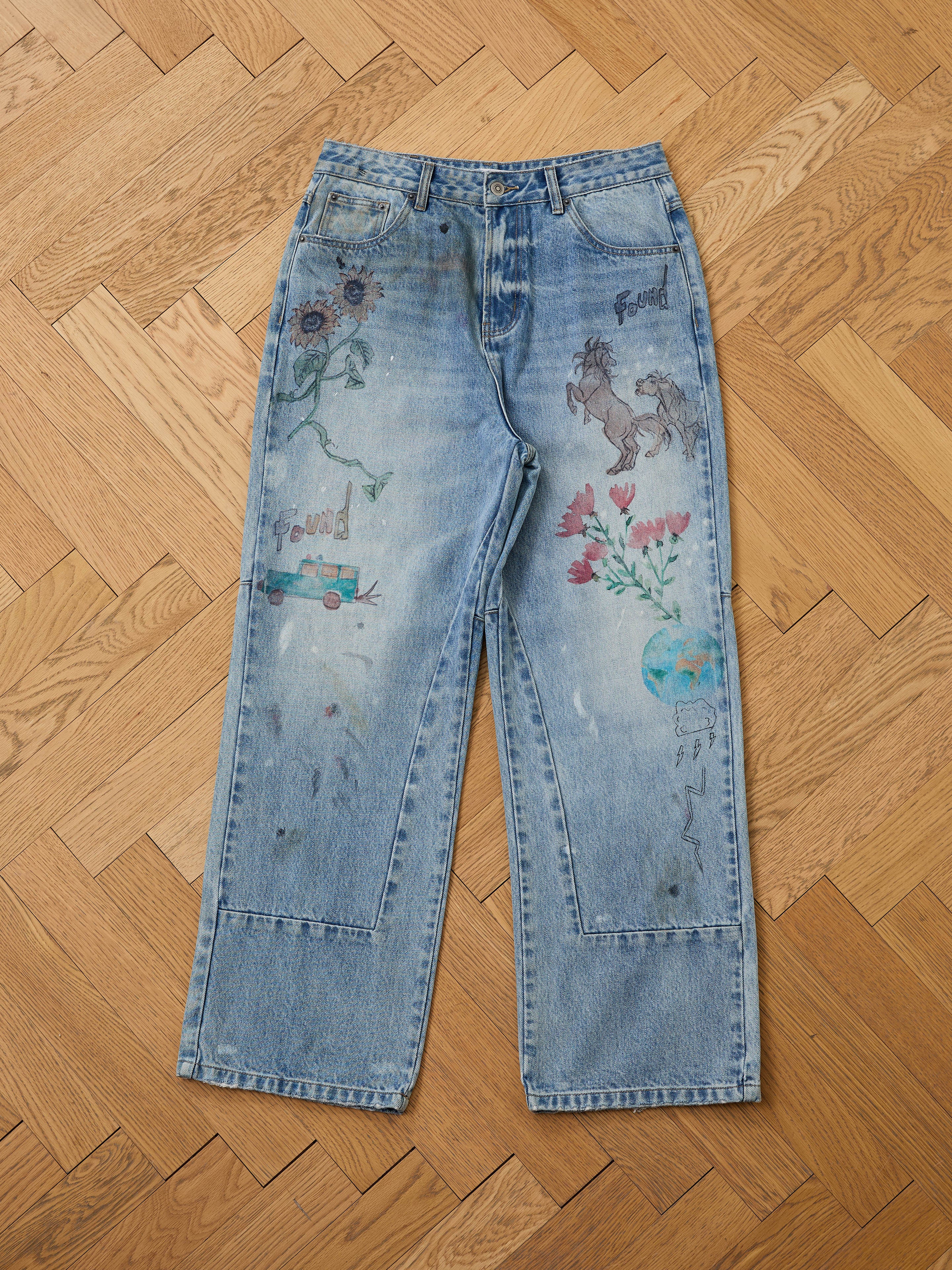 FOUND Hand Art Jeans in light blue denim feature hand-drawn prints like flowers, a horse, a planet, and the word "found." Shown laid flat on a wooden parquet floor.