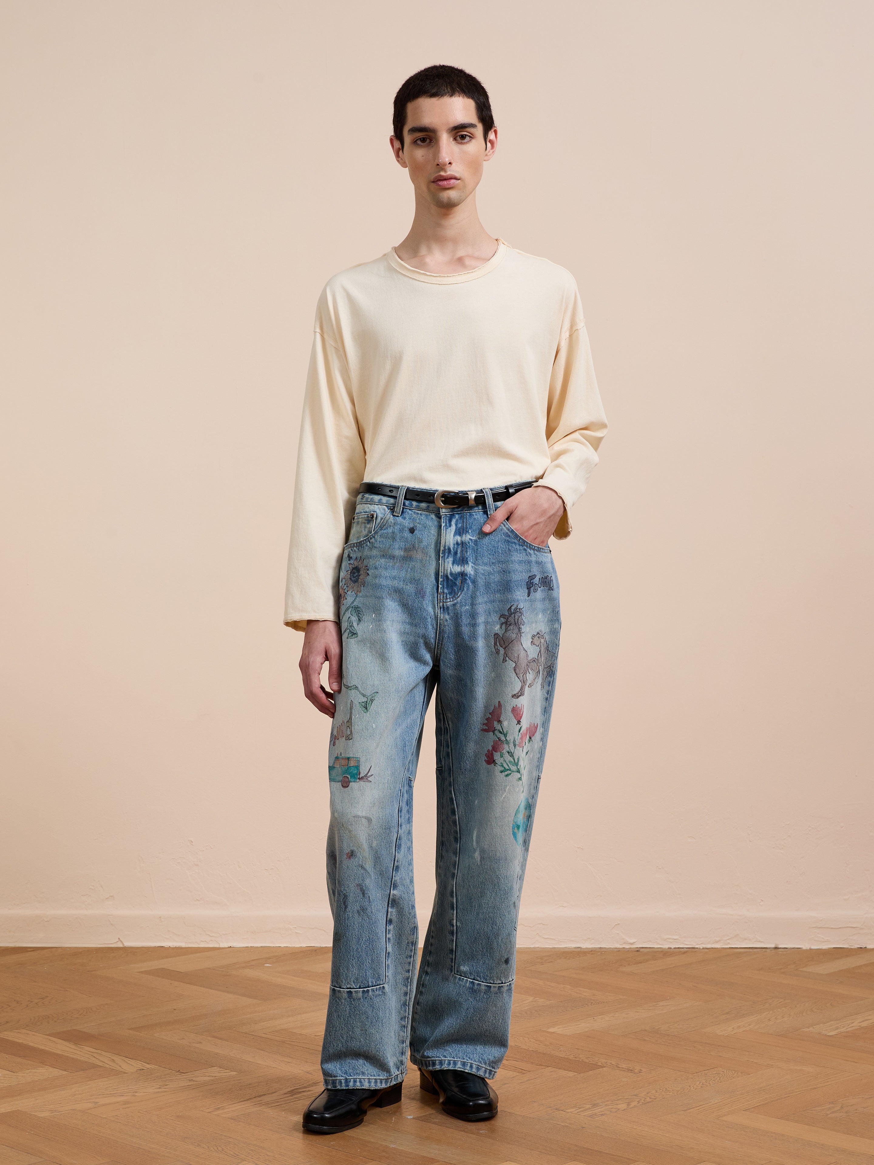 A person stands indoors on a wooden floor, wearing FOUND Hand Art Jeans with original hand-drawn artwork, a cream long-sleeve shirt, and black shoes against a plain background.